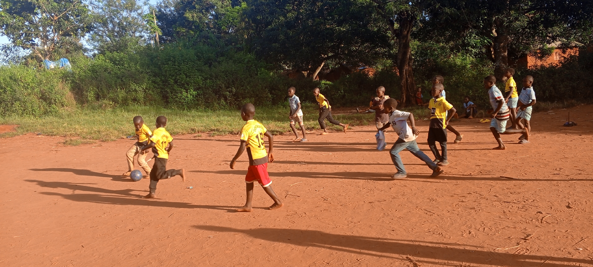 Ngalula, Democratic Republic of the Congo. A group of children playing soccer on a dirt field. The children are of varying ages and are wearing a mix of clothing including some wearing yellow soccer jerseys and some wearing casual clothing. Some children are barefoot. A soccer ball is visible in the foreground. In the background is dense green vegetation. The scene is illuminated by sunlight, casting shadows on the ground.