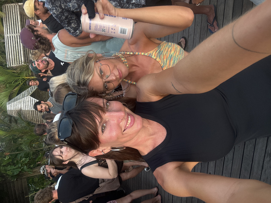 This image captures a crowded outdoor social gathering at a venue in Uluwatu, Indonesia. Two women are prominent in the foreground, posing for a selfie. The woman on the left, closer to the camera, has shoulder-length brown hair with bangs, a nose piercing, and large silver hoop earrings; she wears a black sleeveless top, with dark sunglasses resting on her head. The woman on the right has blonde hair, wears clear-rimmed square eyeglasses with an attached chain, and a multi-colored (orange, yellow, green, light blue) knitted halter-style crop top. She holds a light pink aluminum can, labeled "EVERYDAY WEEKEND," in her raised right hand. Both women are smiling towards the camera.
The midground and background feature a diverse group of approximately twenty additional individuals, mostly young adults, standing and interacting. They are dressed casually in t-shirts, tank tops, and caps. A man in a black t-shirt is visible smiling in the far background, while another man wears a light blue tank top and a maroon baseball cap in the mid-background. The ground surface is composed of dark wooden decking. Tropical foliage and a distinctive angular structure made of light-colored wooden slats are visible in the background, suggesting an open-air setting. The scene is illuminated by natural light.