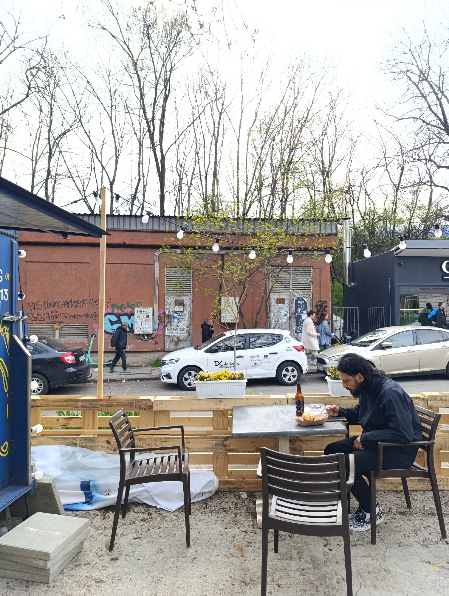The image captures an outdoor dining area in Sofia, Bulgaria, evident from the Cyrillic script on the graffiti and vehicles. A man with long dark hair and a beard, dressed in a dark jacket and pants, is seated at a gray metal table eating from a cardboard container. On the table, there is also a bottle of beer. He sits in a dark brown chair with horizontal slats. Another similar chair faces away from the camera, positioned on the opposite side of the table. The dining space is bordered by a makeshift fence constructed from wooden pallets. The ground is a mix of concrete and exposed earth.
Behind the dining area, there is a street with parked cars: a black sedan, a white compact car labeled "Delivery," and a tan-colored sedan. Pedestrians are visible on the sidewalk. The backdrop features a low, reddish-brown building with graffiti on its walls and a black building. Bare trees dominate the skyline. String lights hang overhead, adding to the ambiance.