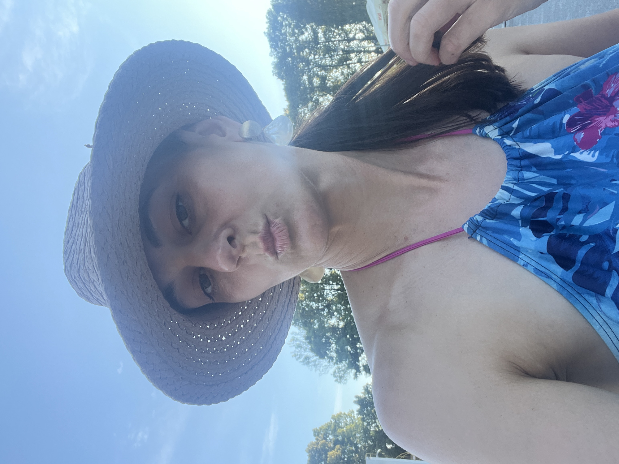 A woman wearing a straw hat and a blue floral bikini is taking a selfie outdoors under a clear blue sky. She is pouting her lips and holding a strand of her dark hair with her right hand. She is wearing large, pearlescent earrings. Behind her, trees and a white tent can be seen.