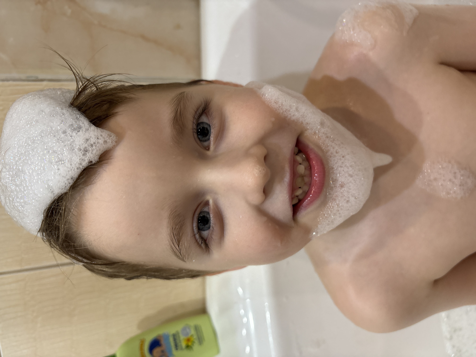 A close-up, rotated image shows a young child in a bathtub covered in bubbles. The child has light skin, blue eyes, and light brown hair, some of which is covered in white bubbles. The child is smiling, showing their teeth. White bubbles are present on the child's face, head, and shoulder. A green bottle is partially visible in the lower left corner of the frame. The background appears to be a tiled wall. The image was taken in a bathroom in the city of Chaikovsky, Russia.