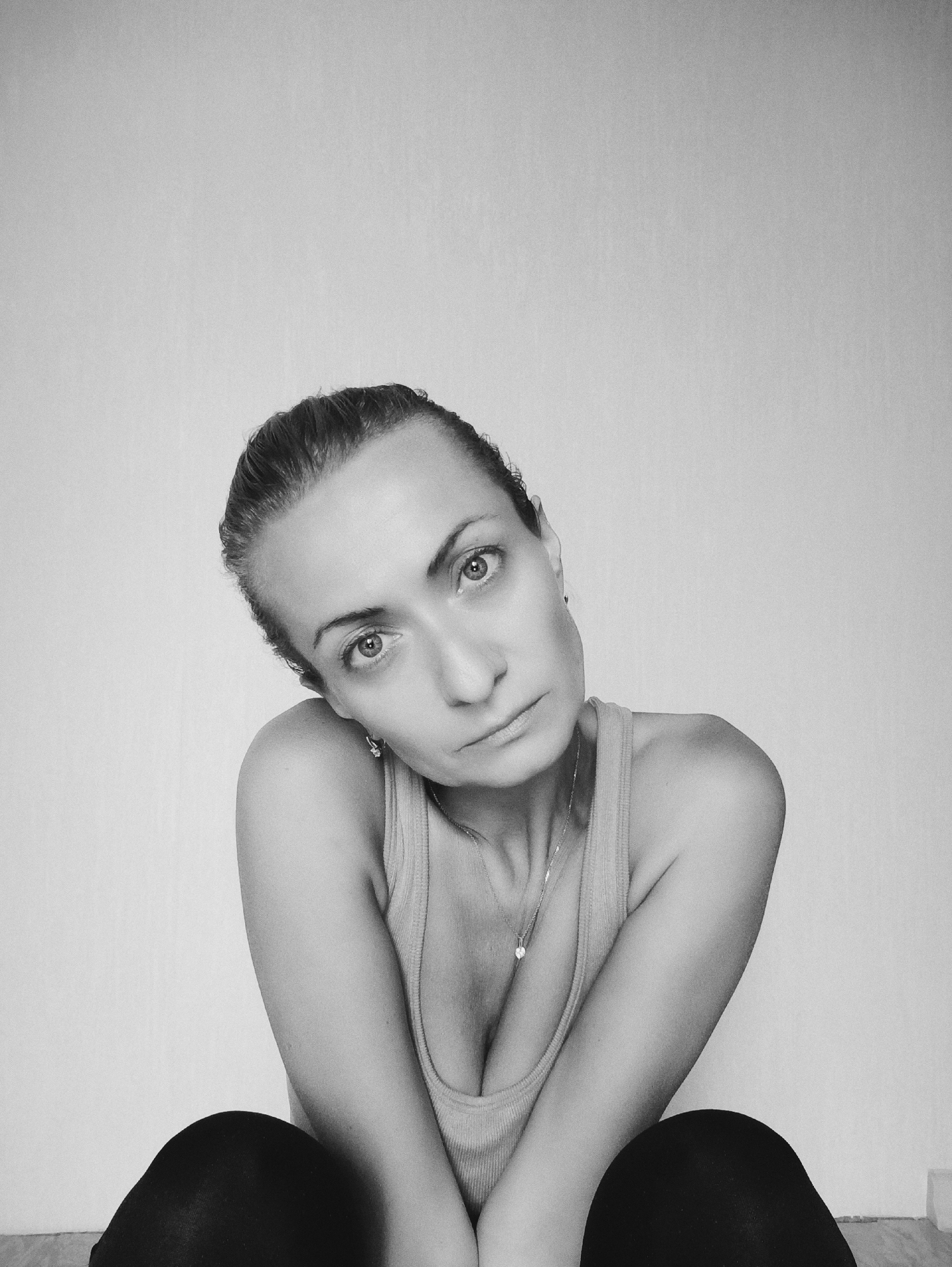 A black and white, medium shot, selfie shows a woman with her head tilted to her right. She has her arms crossed and is looking directly at the camera. She is wearing a tank top and dark leggings. The background is a plain, light-colored wall. The woman is seated on the floor, with her knees bent. The image appears to have been taken indoors, in what seems to be a minimalist apartment in Moscow, Russia.