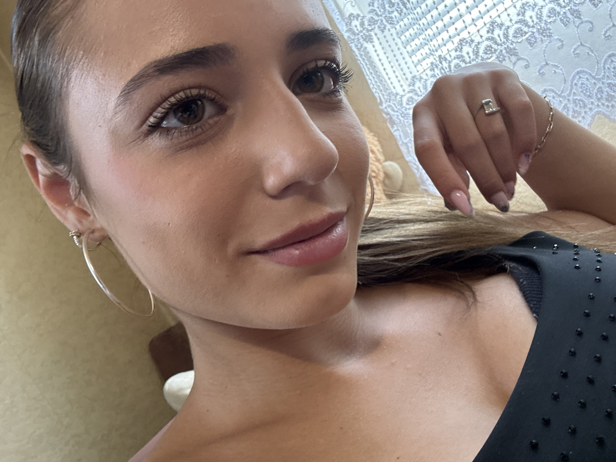 The image shows a close-up, slightly tilted selfie of a young woman. She has dark brown eyes, full lips, and dark hair. She is wearing a black top adorned with small dark studs. On her left hand, she wears a ring on her ring finger and a bracelet on her wrist. She also has hoop earrings in her ears. The background is softly blurred, with hints of a patterned curtain and what appears to be a textured wall. The lighting is natural and highlights her facial features. The scene does not contain any specific markers or landmarks that would allow for the identification of a precise location within the city of Pernik, Bulgaria.