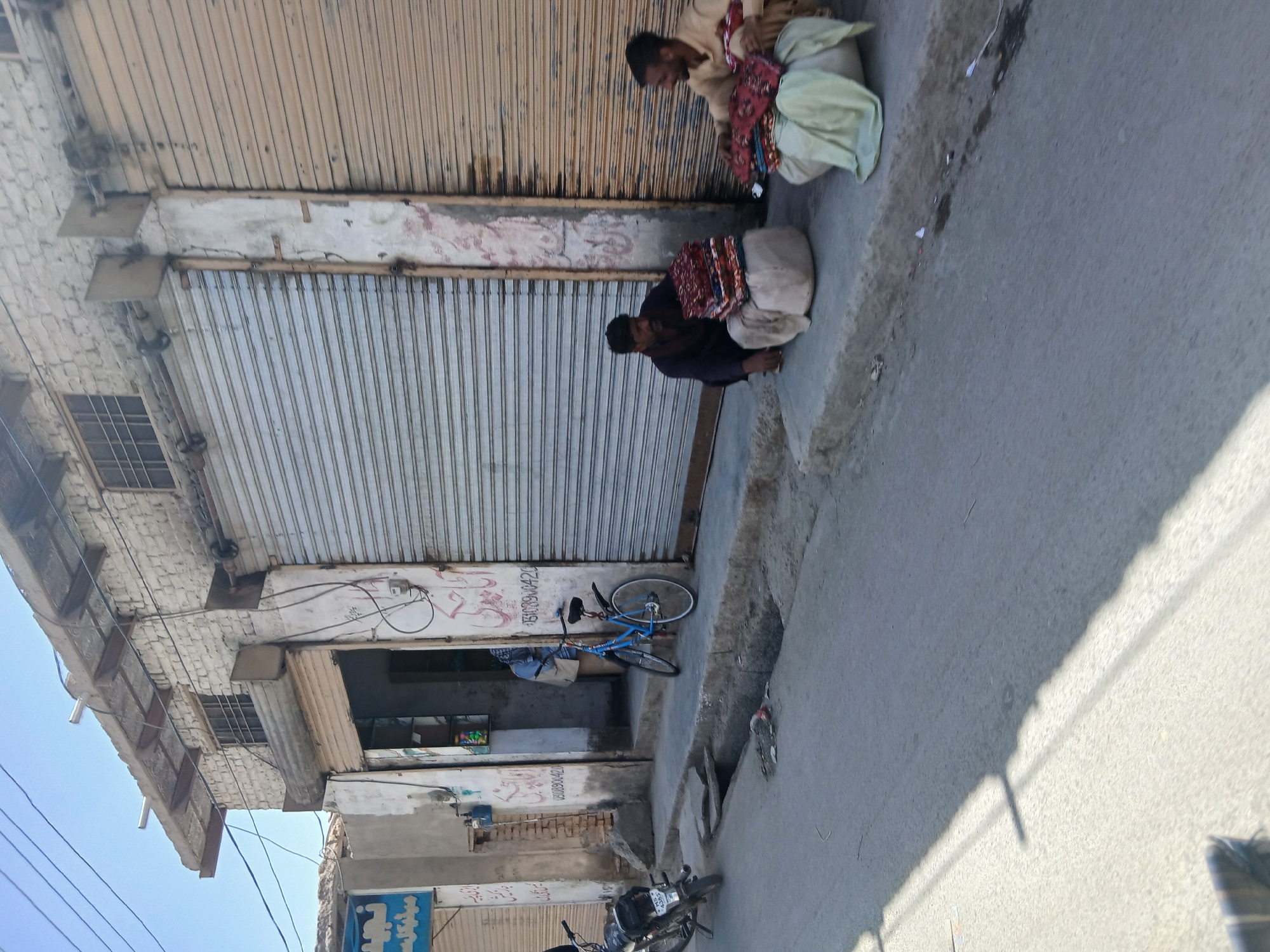 The image depicts a street scene in Quetta City Tehsil, Pakistan. Two men are sitting on the sidewalk in front of a closed storefront with a corrugated metal roll-up door. They appear to be sorting through or displaying fabrics. A blue bicycle is leaning against the wall next to a partially open shop, and a motorcycle is parked further down the street. The buildings show signs of age and wear.