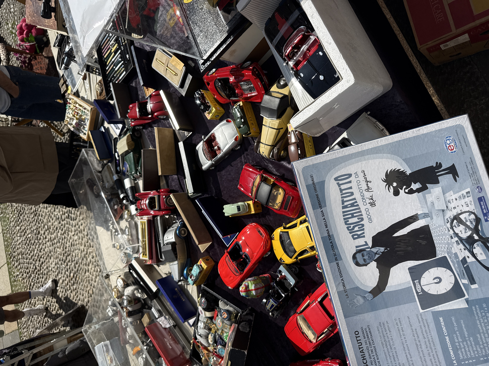 The image shows a table at an outdoor market. On the table are a variety of items, including model cars, pens in boxes, and a boxed board game called "Il Rischiatutto." People are browsing the items on the table. The market appears to be on a paved street.