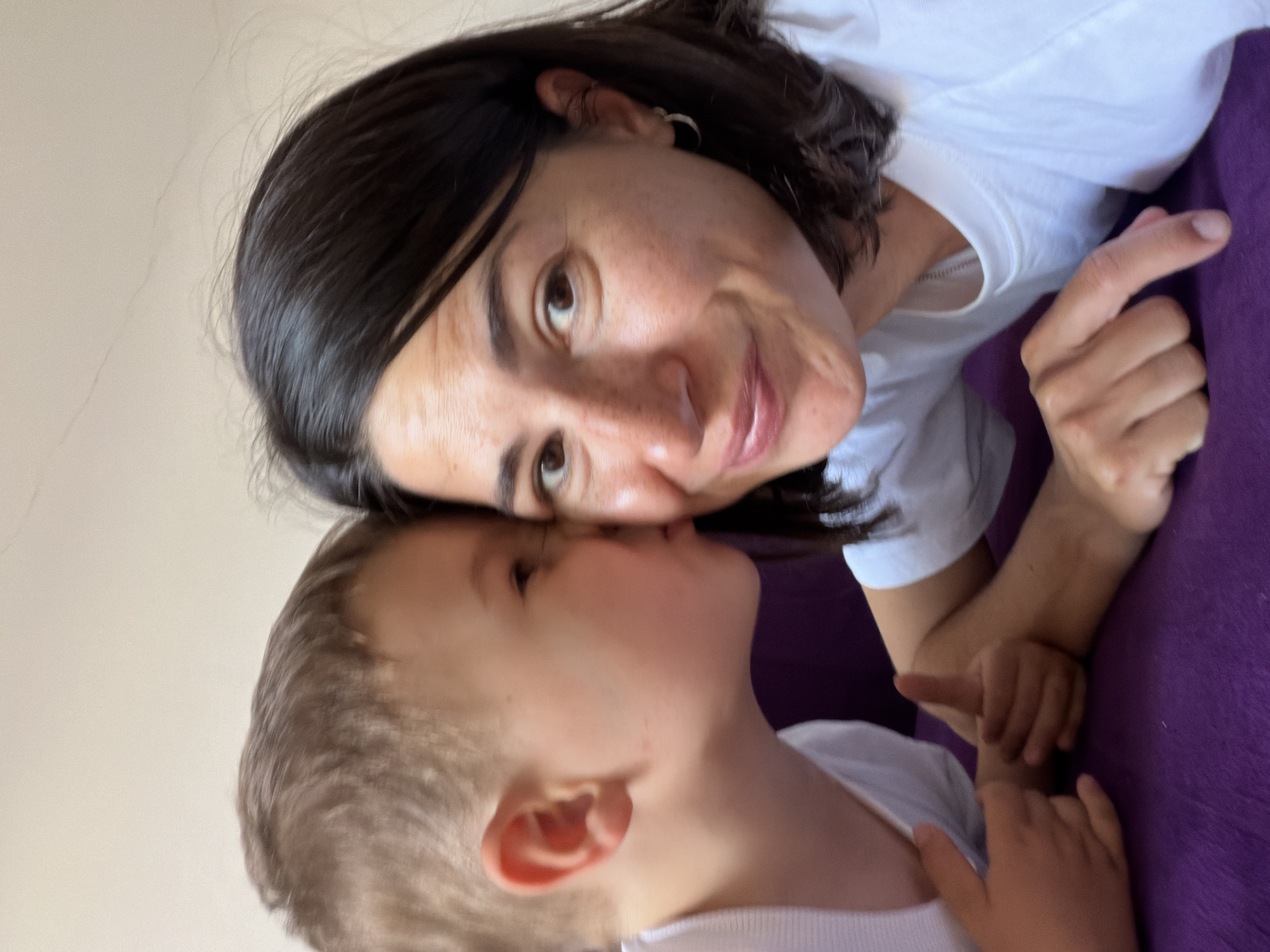 This close-up image captures an intimate moment between an adult female and a young boy, reportedly situated in Budva, Montenegro. The two subjects are positioned closely, with the boy, who has short light brown hair and wears a white t-shirt, giving a kiss to the adult female's right cheek. The female, with dark, shoulder-length hair, brown eyes, and visible freckles, looks towards the viewer with a slight smile and puckered lips. She also wears a white t-shirt and a small silver hoop earring in her visible left ear. Her right index finger is extended, pointing downwards towards a deep purple textile surface that occupies the lower portion of the frame, likely a couch or bed. The boy's right hand is visible, making a 'thumbs up' gesture, while his left arm and the woman's left hand appear to be intertwined or resting together on the purple surface. The background consists of a plain, light-colored wall.