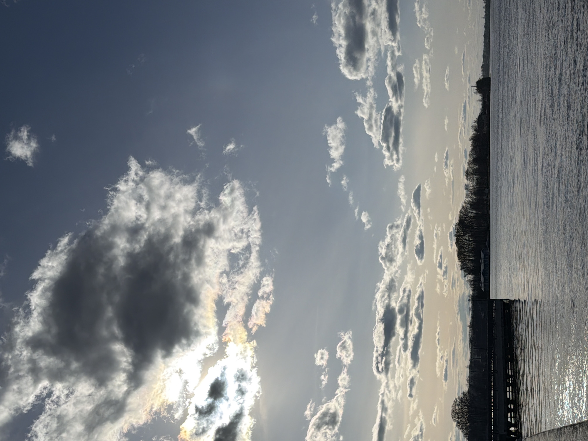 The image depicts a sky with cumulus clouds, some obscuring the sun. The lower portion of the image shows a body of water, possibly a river or lake, reflecting sunlight. A line of trees is visible along the shoreline, along with a structure resembling a pier or embankment. There is no evidence to confirm the specific location in Votkinsk, Russia.