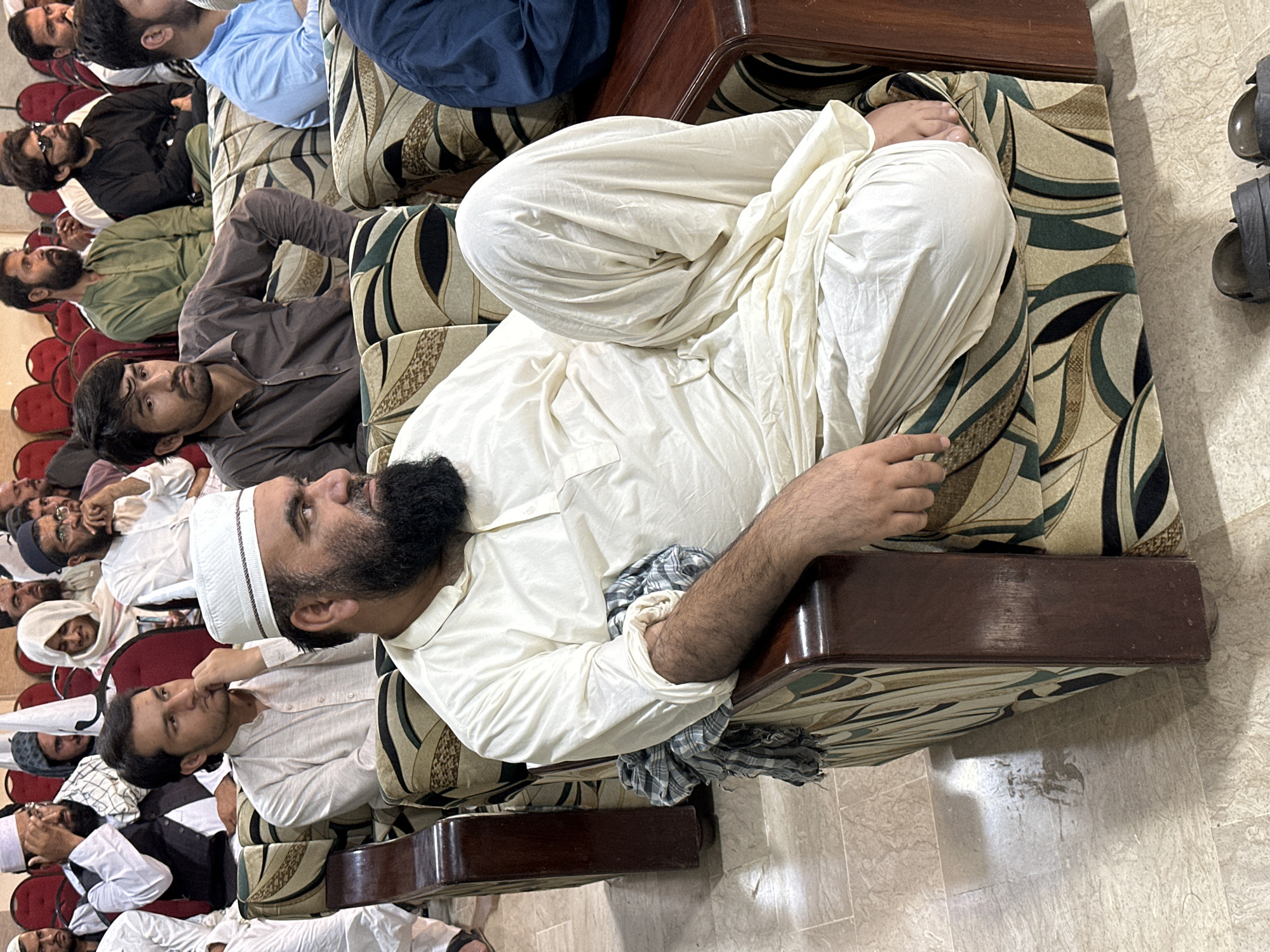In a room within Quetta City Tehsil, Pakistan, a group of men are seated on upholstered chairs and red plastic chairs. The focal point is a man in traditional white attire and a white cap, reclining on a patterned couch. He has a beard and is positioned among the seated individuals. Several other men wear similar traditional clothing and caps, while some wear Western attire. The seating arrangement appears to be in rows, suggesting a gathering or meeting. The room's interior features a tiled floor and patterned upholstery on the furniture. A pair of shoes is visible on the floor, to the right.