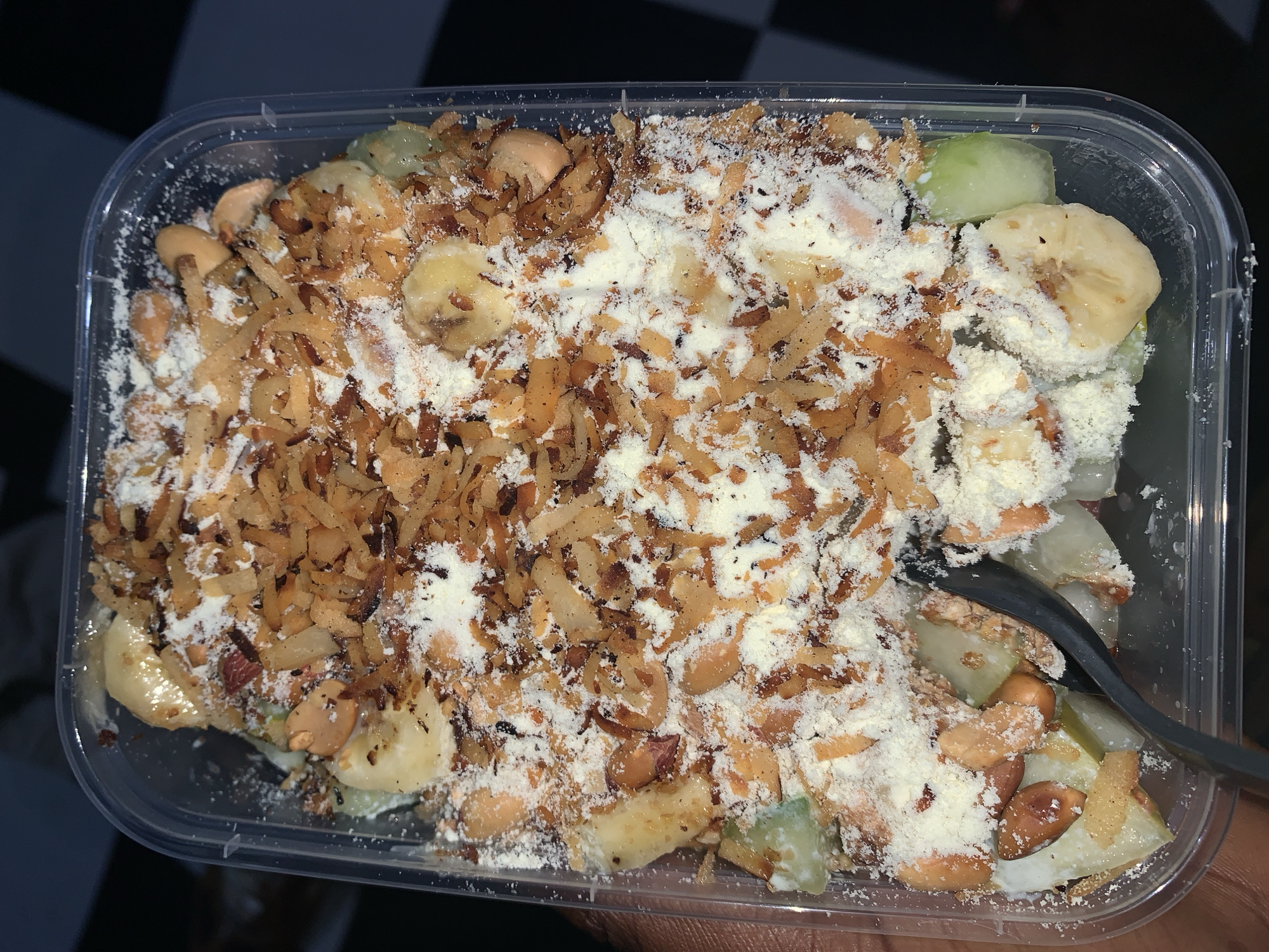 The image displays a clear, rectangular plastic container filled with a multi-textured food item, viewed from directly above. The contents appear to be a fruit salad or dessert.
The base layer consists of chopped fruits, including distinct slices of banana and pieces of a light green fruit, possibly melon or cucumber. Mixed throughout the fruit are whole peanuts, some still retaining their reddish-brown skins, while others appear blanched.
A substantial layer of toppings covers the fruits and nuts. This includes a dense scattering of toasted, golden-brown, shredded flakes, likely coconut or a similar crunchy ingredient. Over this layer and directly on other components, a thick and uneven dusting of white powder is spread, which could be powdered sugar, milk powder, or a fine spice blend.
On the right side of the container, a dark-colored spoon or utensil is partially visible, inserted into the food, suggesting it is ready for consumption or is currently being eaten.
The background is predominantly dark, with a black and white checkered pattern visible in the upper left quadrant, indicating an indoor setting such as a tiled floor or a patterned surface. The lighting appears artificial. There are no people visible, nor any discernible text on the container or in the background. The dish presents as a prepared snack or dessert from Rafin Par, Nigeria.