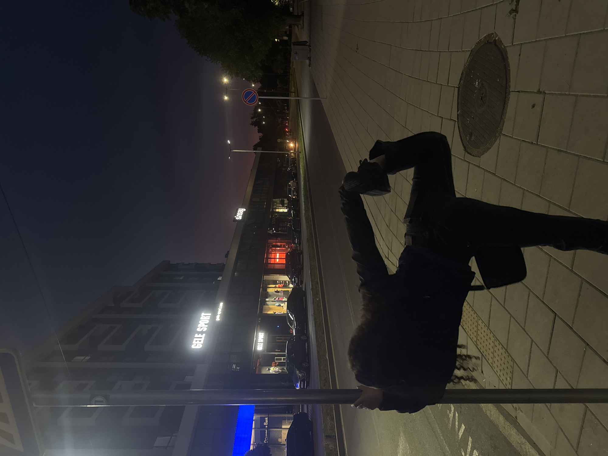 In a nighttime urban scene, a person with dark, curly hair is shown from behind, performing a pose against a metal pole. They are dressed in dark clothing, including a jacket and pants, and appear to be balancing with one leg extended. The setting is a sidewalk paved with rectangular tiles, adjacent to a street. In the background, a multi-story building with illuminated storefronts is visible, featuring signs for "GELE SPORT" and "sinsay". Cars are parked along the street and moving along it. Streetlights illuminate the sidewalk and road. The sky is a deep twilight blue with a hint of purple near the horizon. A traffic sign with a red circle and a diagonal bar is also present on the sidewalk.