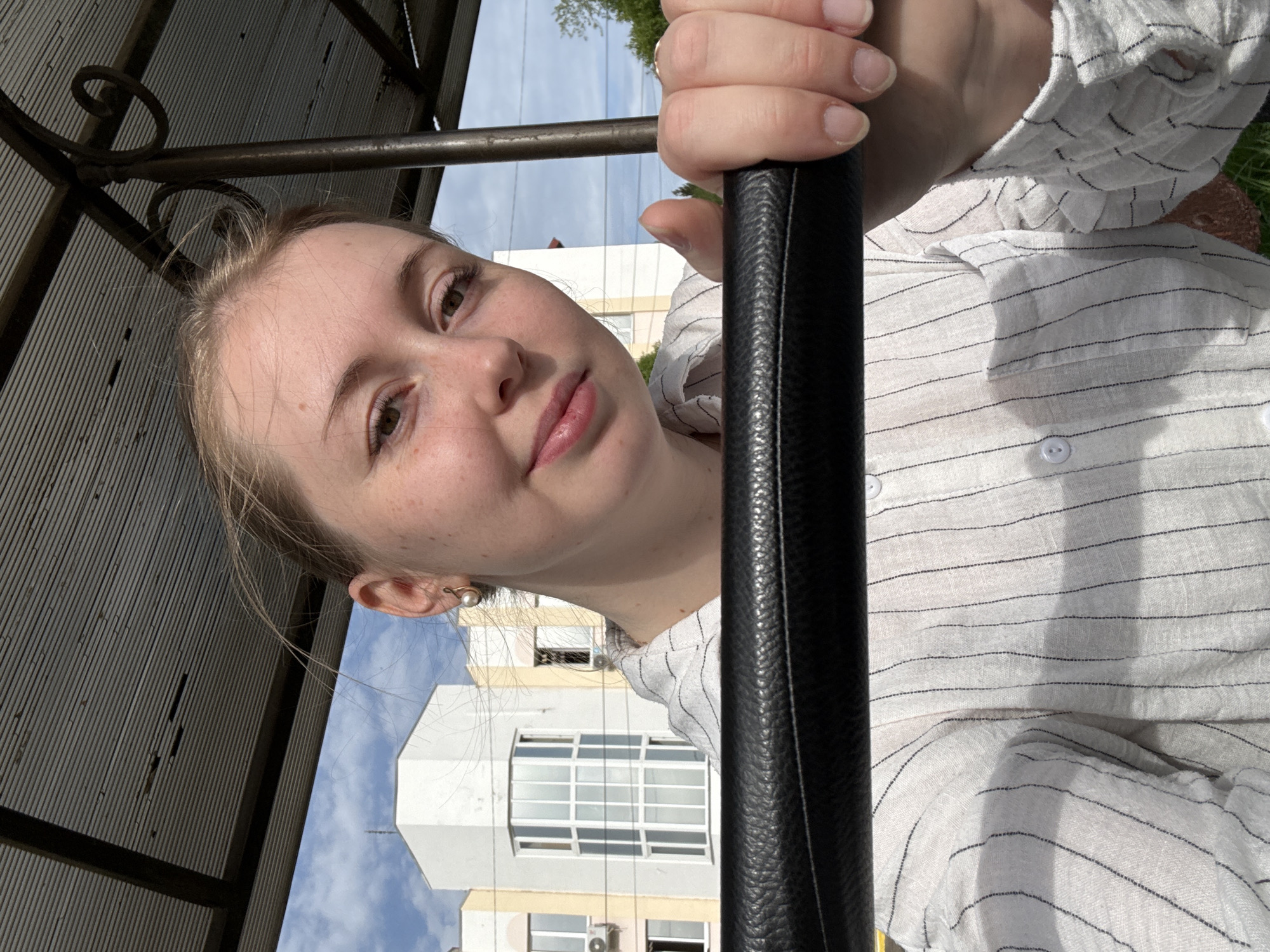 A close-up, slightly angled shot features a fair-skinned person with light brown hair pulled back. They are wearing a white, vertically striped shirt and are holding onto a black, cylindrical object, possibly a handle. The background reveals a multi-story building with several windows. Above the building is a blue sky with light clouds. The person appears to be under some type of structure. The location is in an urban area, Unknown, Unknown.
