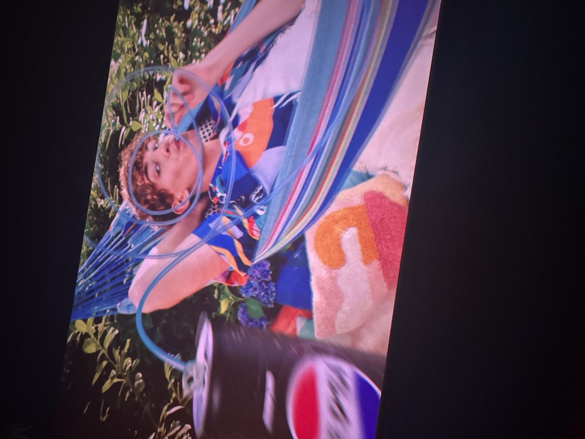 An image, captured at an angle with a black border on the left and right, shows a young person with curly brown hair relaxing in a blue string hammock outdoors. The person is wearing a vibrant, geometrically patterned shirt in shades of blue, orange, white, and black. A blue tube or wire wraps around their head and body, possibly indicating headphones or a hydration system.
Behind the person, lush green foliage suggests a garden or natural setting. A colorful striped fabric or blanket in blues, pinks, whites, greens, yellows, and oranges is draped over the hammock and the person's lower body. A cluster of blue flowers, possibly hydrangeas, is visible near the person's torso.
In the foreground, a beverage can, out of focus and partially obscured, is visible. Its red, white, and blue circular logo strongly resembles that of Pepsi. The scene appears brightly lit, indicating it is daytime. The person's expression is relaxed, and they are looking upwards or slightly to the side. The overall impression is one of leisure and summer relaxation.