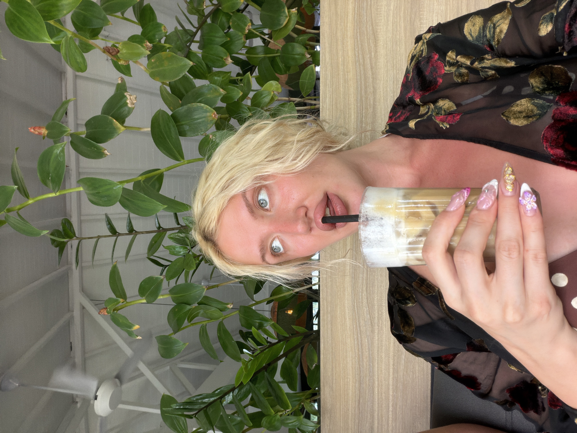 In North Kuta, Indonesia, a Caucasian woman with blonde, wavy hair and striking blue eyes is captured in a medium close-up shot, holding a tall glass of iced coffee with a dark straw. She is wearing a sheer black kimono-style robe adorned with a dark floral pattern featuring red and gold accents. Her makeup is visible, with emphasis on her lips and eyes. Her nails are long and elaborately decorated with rhinestones and intricate designs. The background features lush green plants, suggesting an outdoor or semi-outdoor setting, possibly a cafe or restaurant. A ceiling fan is visible in the upper left of the image, and a wooden panel forms the backdrop behind the woman.