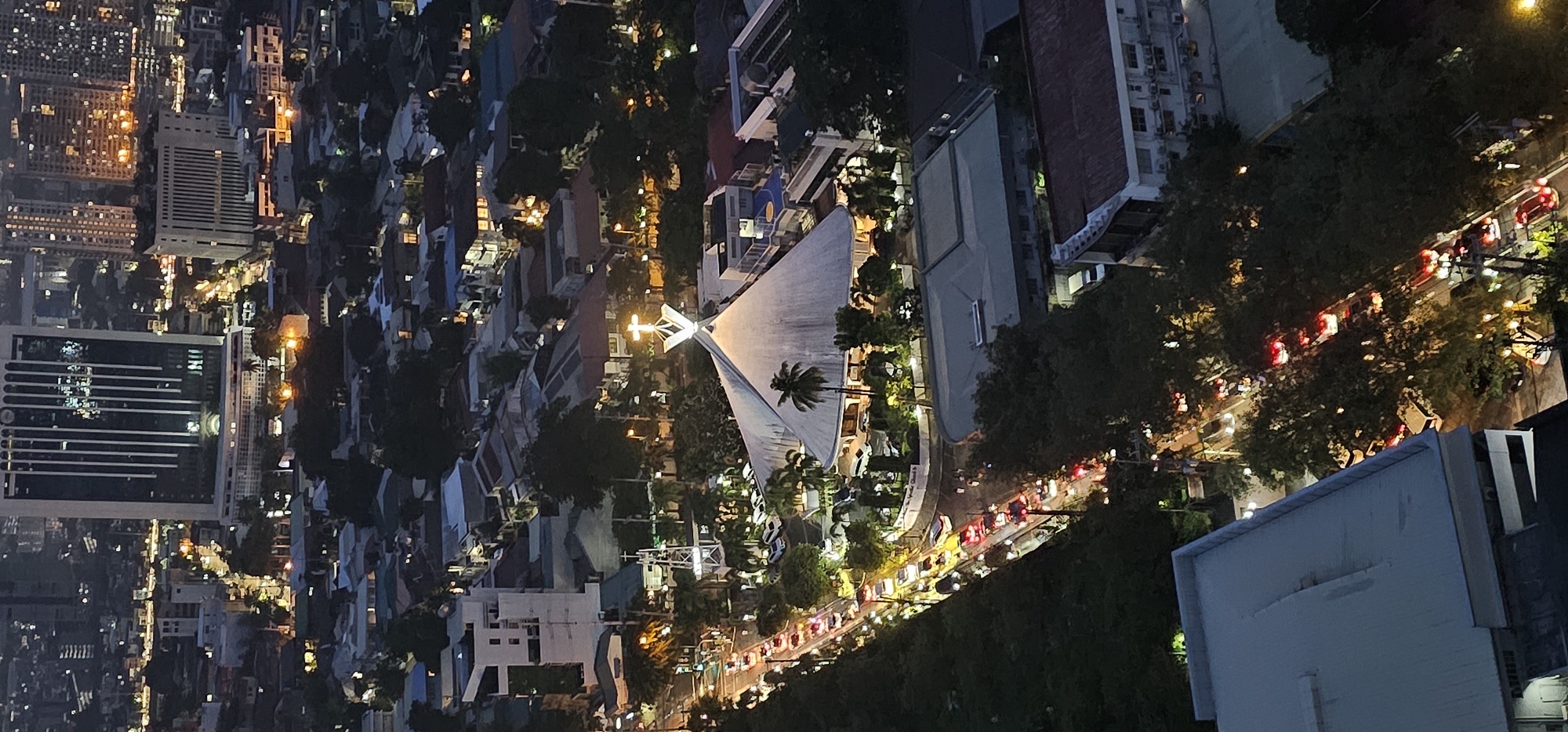 This is an aerial, nighttime photograph of a city scene in Makati, Philippines. The focus is on a large, modern church with a distinctive white, conical roof topped with a glowing cross. The church appears to be located on a wide street, which is congested with traffic. Numerous cars, their headlights and taillights visible as streaks of light, are lined up along the road.
Surrounding the church and the street are various buildings, including residential houses with their roofs illuminated, and taller, more modern structures, one of which is a prominent skyscraper with vertical lighting. Lush green trees are interspersed throughout the landscape, some lining the street and others surrounding the church. Streetlights are visible, casting a warm glow on the scene. The overall impression is of a bustling urban environment at night.