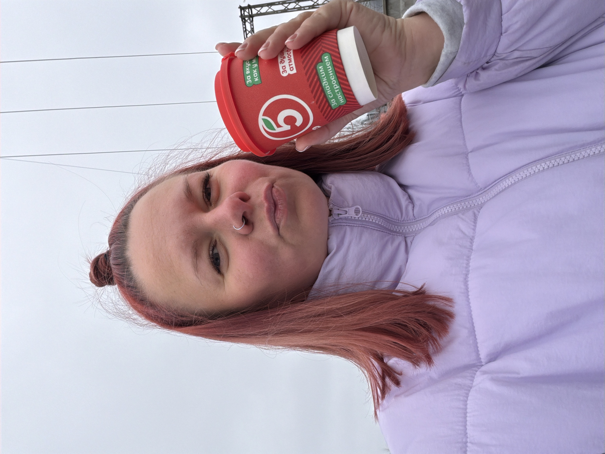 A red-haired woman with a nose ring holds a red and white coffee cup with Russian writing. She is wearing a light purple puffer jacket. Overhead wires can be seen in the background, suggesting an urban environment. The image was taken outdoors in Chaikovsky, Russia.