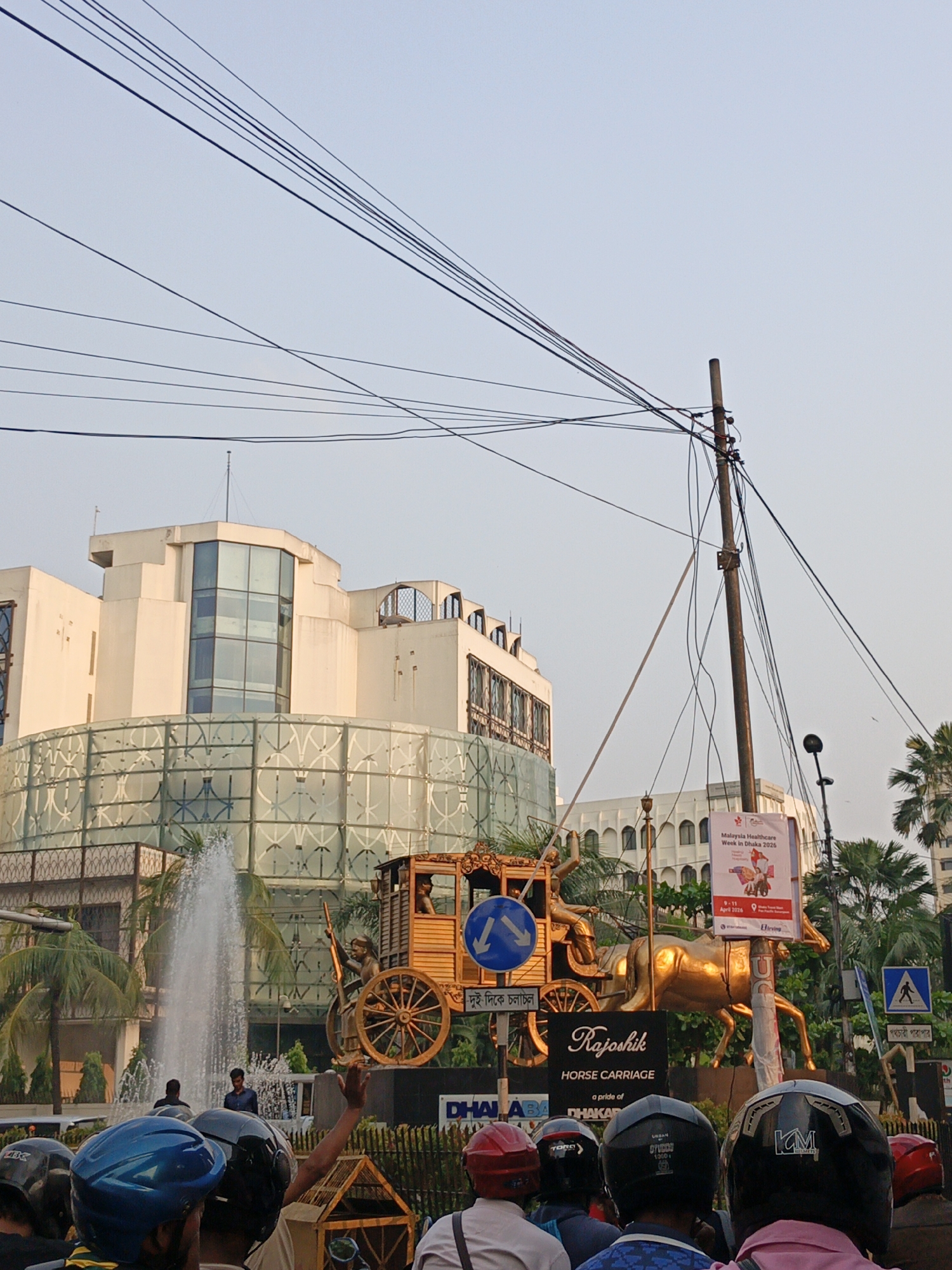 The image captures a street scene in Dhaka, Bangladesh, featuring a golden horse-drawn carriage statue labeled "Rajoshik Horse Carriage" surrounded by a crowd of people wearing helmets, suggesting they are likely motorcyclists or scooter riders. In the background, there is a modern building with a curved, glass-enclosed facade, and a fountain in front of it. A street sign indicates a pedestrian crossing. Power lines crisscross overhead, and a utility pole is visible with a sign advertising "Malaysia Healthcare Week in Dhaka 2026."