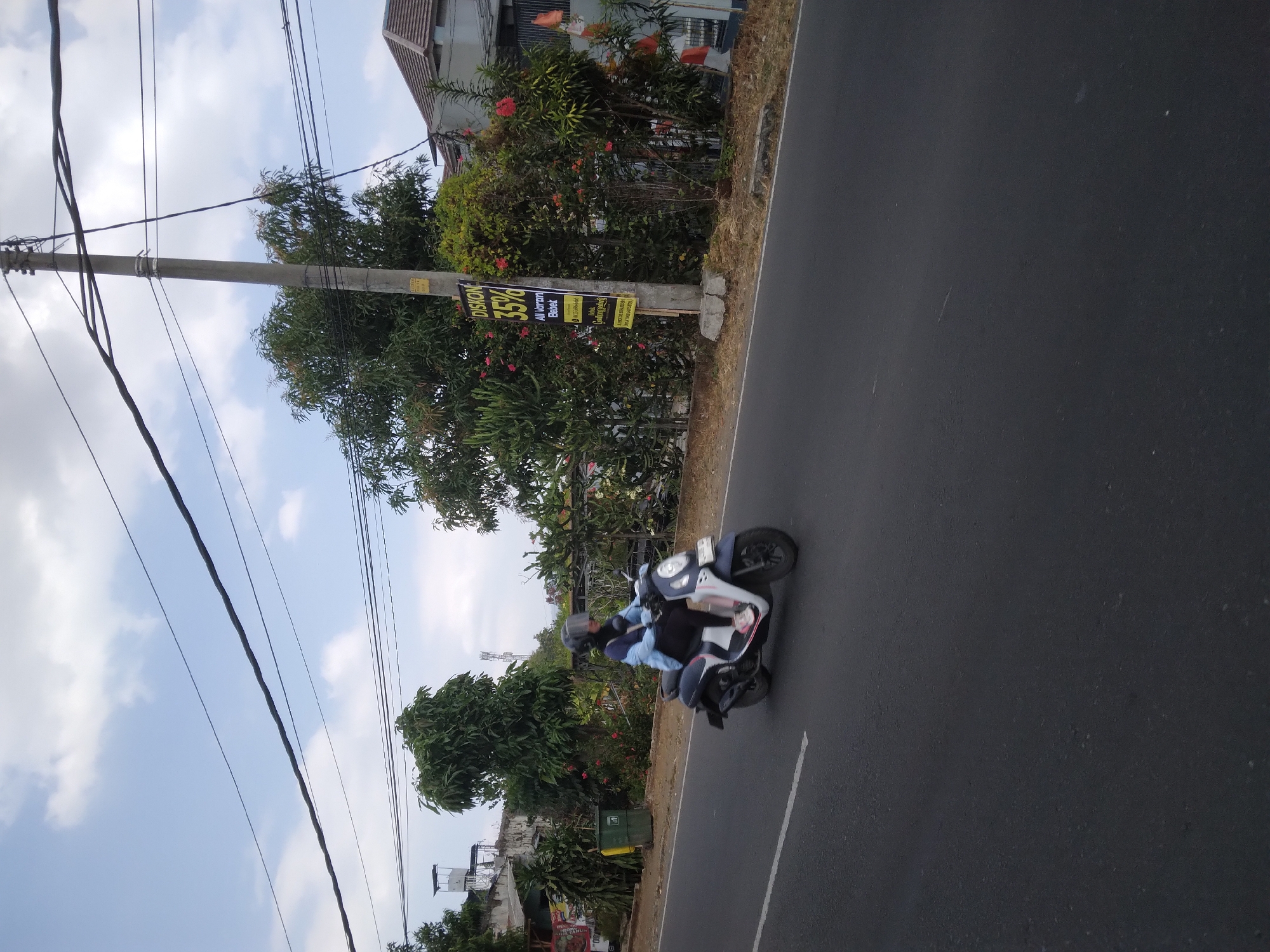 A full shot shows a person riding a scooter on a paved road in Berkoh, Indonesia. The person is wearing a blue jacket, black pants, and a helmet. The scooter is dark blue and white. The road is asphalt with a white line on the left. Trees and bushes are on the right side of the road. A utility pole with wires is also visible. A sign on the utility pole advertises a 35% discount. The sky is blue with some clouds.