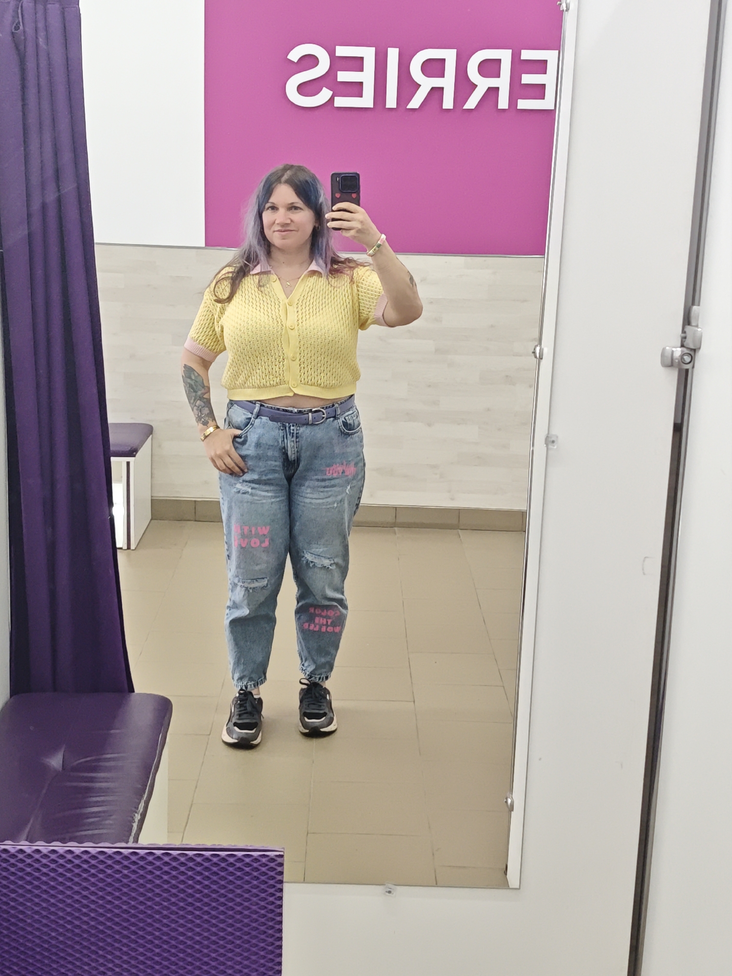 A woman is taking a selfie in a changing room. She has blue-tinted hair and is wearing a yellow cardigan, jeans with pink lettering, and athletic shoes. The changing room has a purple bench and curtain, a white wall with "BERRIES" spelled backwards in pink letters, and a tiled wall above the floor. Based on the lettering, the location is likely a Berries store in Ильинское, Russia.