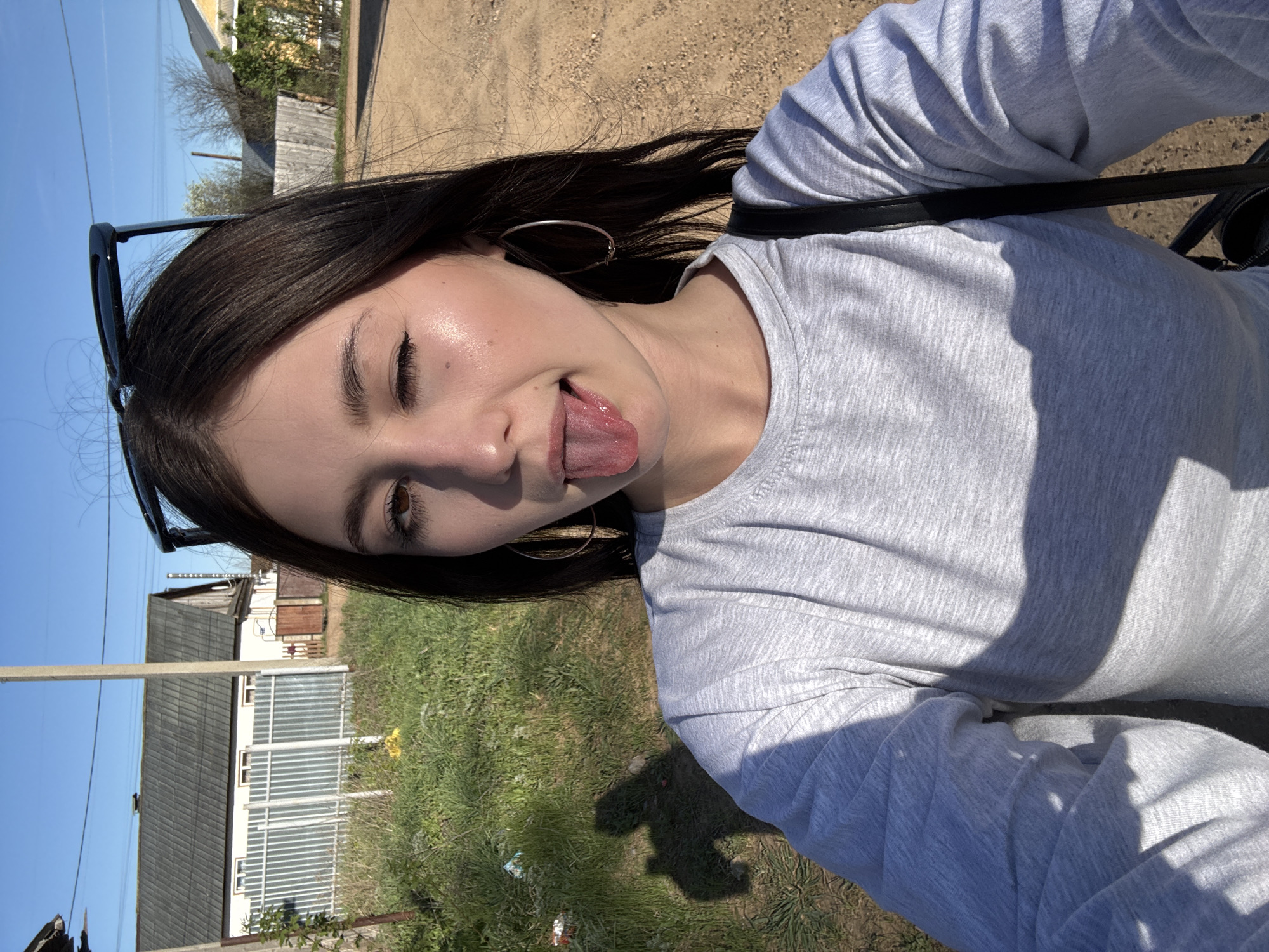 The image is a selfie of a young woman with long, dark hair and fair skin. She is wearing a grey long-sleeved shirt and gold hoop earrings. Sunglasses are perched on top of her head. She is sticking her tongue out and closing one eye.
The background shows a rural or suburban area. There are small houses with corrugated metal roofs and white metal fences. Green grass and patches of dirt are visible. Power lines are present in the sky. The light suggests it is daytime. The location is in the city of Фоки, Russia.