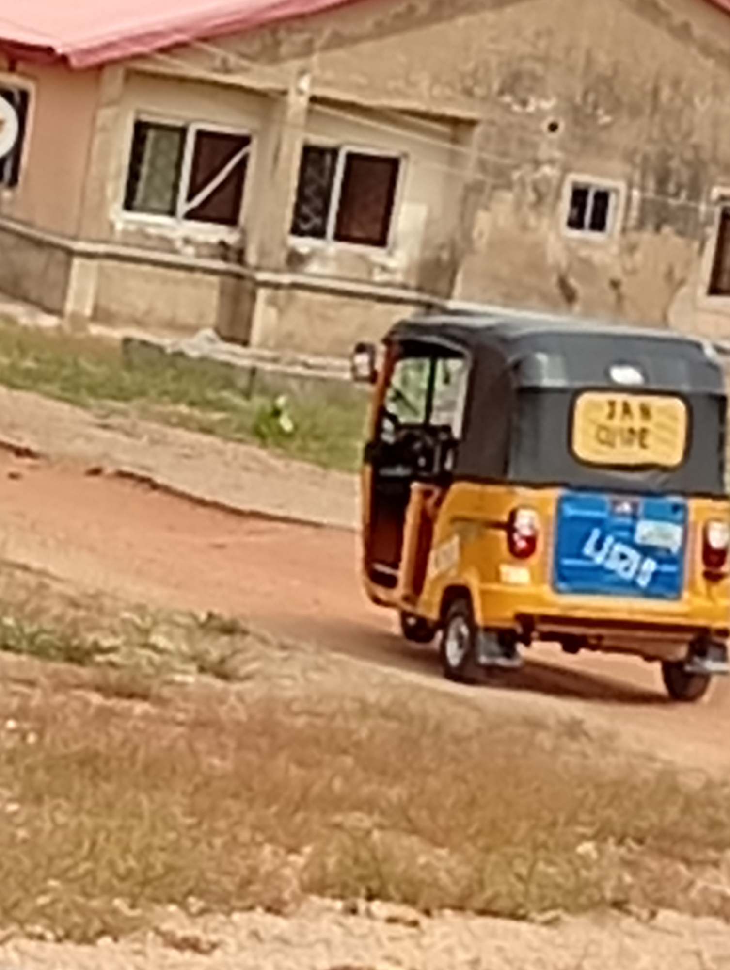 A yellow auto-rickshaw is moving to the right on a dirt road in an urban environment. The auto-rickshaw has a black roof, and a blue panel on the back. The writing "JAN CLIVE" is visible on the back of the roof. Behind the auto-rickshaw is a light-colored building with a red roof and windows. The surrounding area consists of dirt, grass, and sparse vegetation. The exact location within the city of Unknown, Unknown, cannot be determined with certainty from the image.