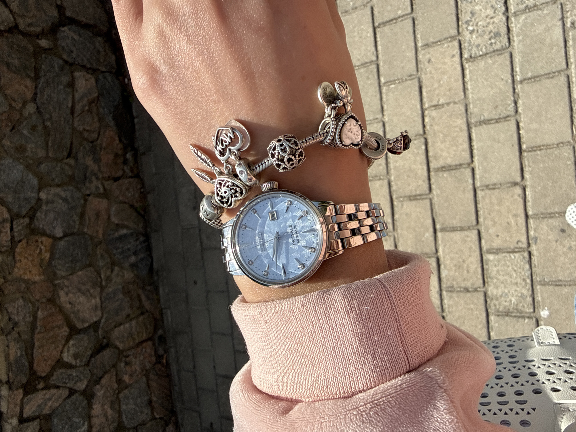 A close-up shot shows a person's left wrist adorned with a silver watch and a Pandora bracelet featuring various charms. The watch, with a light blue face and rose gold accents, is positioned above a stone wall and a paved pathway. The bracelet, also silver, is decorated with charms including hearts, feathers, and a crown. The background consists of a textured stone wall and a light-colored brick pathway. The lighting suggests an outdoor setting. The image does not contain enough information to determine the specific location in Berdychiv, Ukraine.