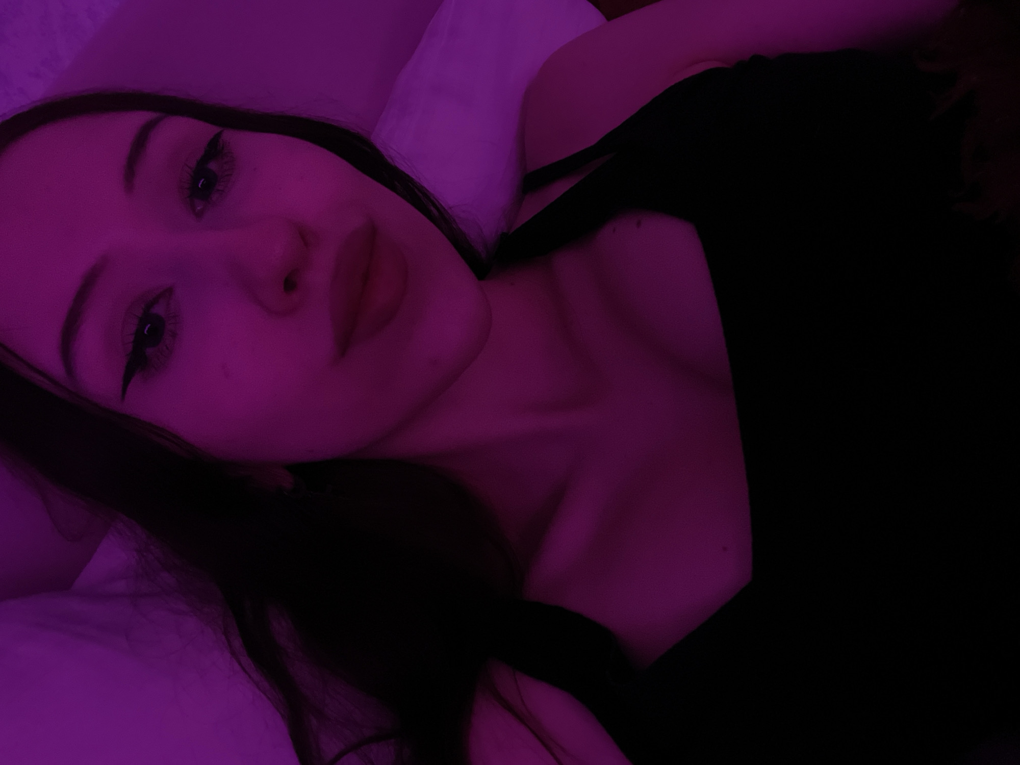 The image is a close-up selfie of a young woman with long, dark hair, lying down. She is wearing a black top. The lighting is predominantly purple, casting a glow on her face and body. Her eyes are open, and she has winged eyeliner. Her lips are full and appear to be painted with a red lipstick. The background is blurred and appears to be a bed or a similar soft surface, also illuminated by the purple light. There are no discernible elements in the image that would suggest a specific location in Votkinsk, Russia, or any other city.