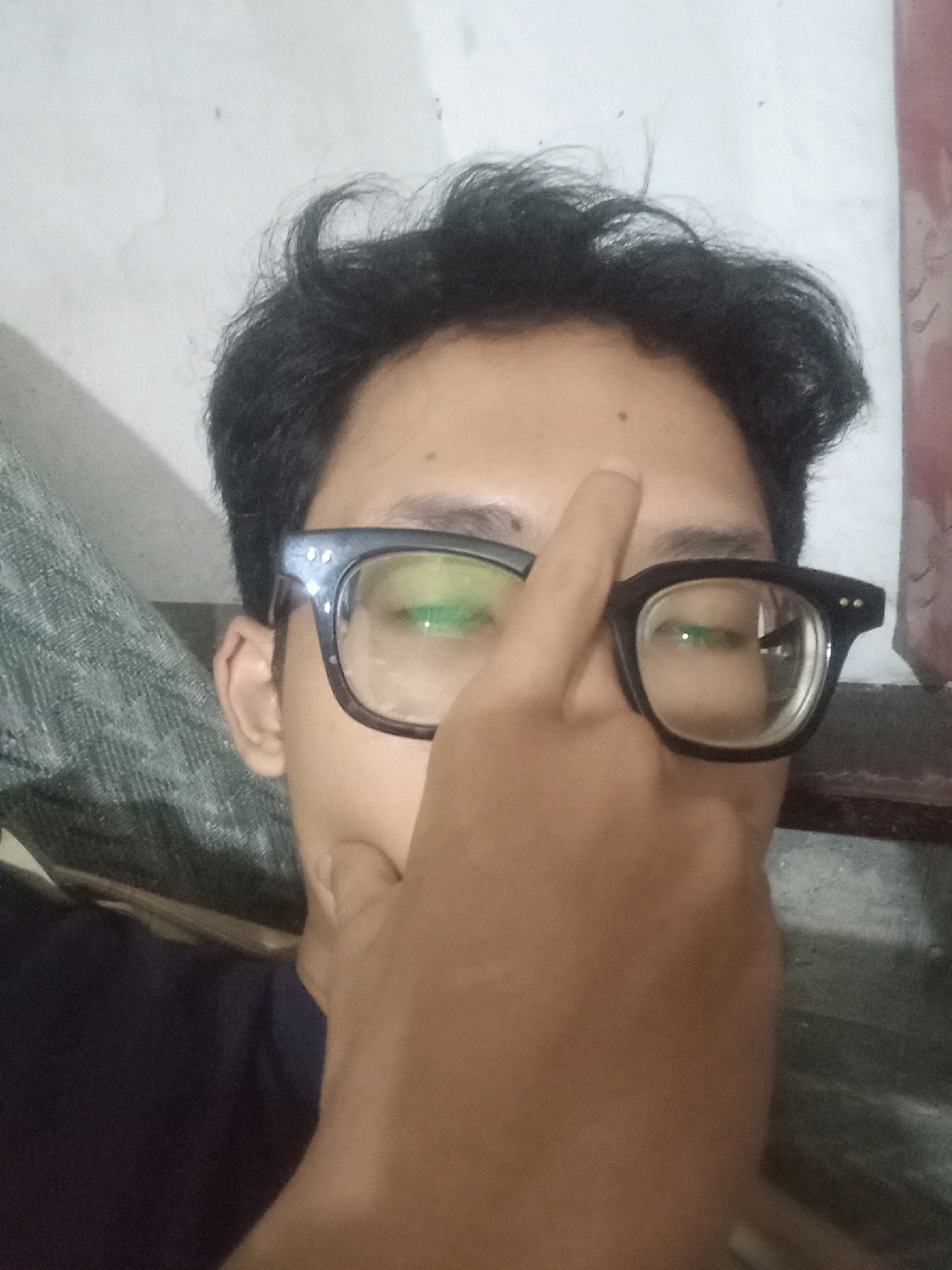 In the city of Cintaratu, Indonesia, a close-up, medium-shot, eye-level image captures a person wearing black-rimmed glasses. The person's right eye is visible through the lens, reflecting a greenish hue. Their right hand is raised, with a finger touching their forehead, partially obscuring their face. The background consists of a light-colored wall, with a portion of a dark fabric to the left and a reddish-brown patterned fabric to the right. The person's dark, messy hair is also visible. The lighting in the image appears to be artificial, casting soft shadows.