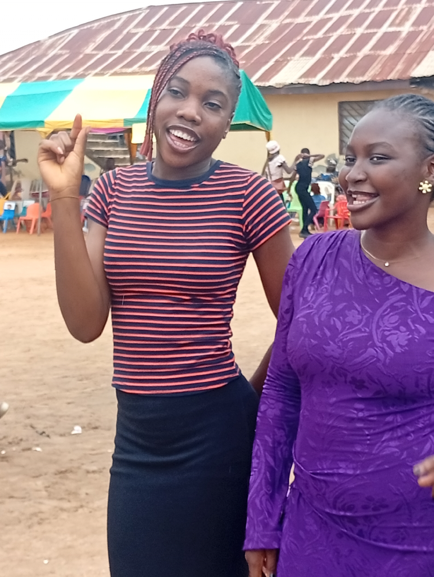 Two young women stand outdoors in front of a building with a corrugated metal roof, partially covered by a yellow and green tent. The woman on the left, with braided hair, wears a striped t-shirt and a black skirt, and raises her right hand with her index finger extended. She appears to be speaking or gesturing. The woman on the right wears a long-sleeved purple dress with a floral pattern and a small pendant necklace. She is smiling and looking to her right. Several other people and colorful chairs are visible in the background, suggesting a gathering or event. The ground is sandy.
