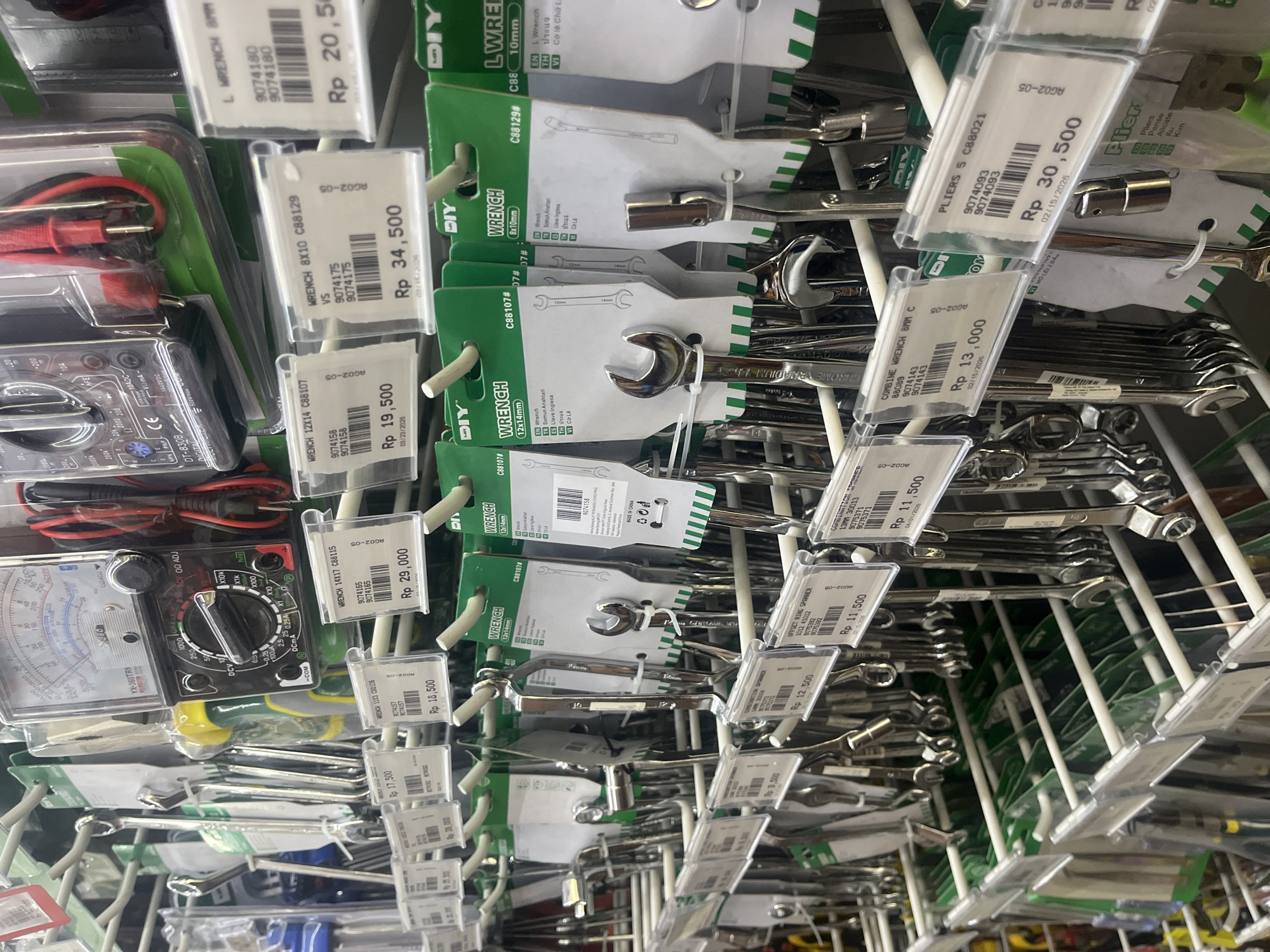The image captures a detailed view of a hardware store's display shelf in West Jakarta, Indonesia, featuring various tools arranged for sale. The products are displayed on white wire racks, hanging at an angle due to the image's rotation.
The primary subjects are numerous hand tools, including a wide assortment of wrenches and pliers, many branded "DIY". Specific items visible with their price tags include an "L WRENCH 8MM" priced at Rp 20,500, a "WRENCH 8X10 C88129" for Rp 34,500, and a "WRENCH 12X14 C88107" for Rp 19,500. Pliers, specifically "PLIERS 5 C88021," are available for Rp 30,500. Other wrenches, such as "COMBINE WRENCH 8MM C" (Rp 13,000) and an "OFFSET RING SPANNER 13MM" (Rp 11,500), are also part of the display.
Beyond wrenches, two multimeters are visible: an analog model "YX-360TRN" and a digital "DT-830B DIGITAL," each accompanied by red and black probes. Each product has a plastic price tag clearly showing its cost in Indonesian Rupiah (Rp), product codes (e.g., 9074180, AG02-05, C88129), and barcodes. Some tags also include dates like "02/15/2026" or "03/23/2026." The packaging for some wrenches features the word "Wrench" in multiple languages, including English, Bahasa Indonesia, Spanish, Thai, and Vietnamese. The overall scene depicts a well-organized retail environment dedicated to hardware and tools.
