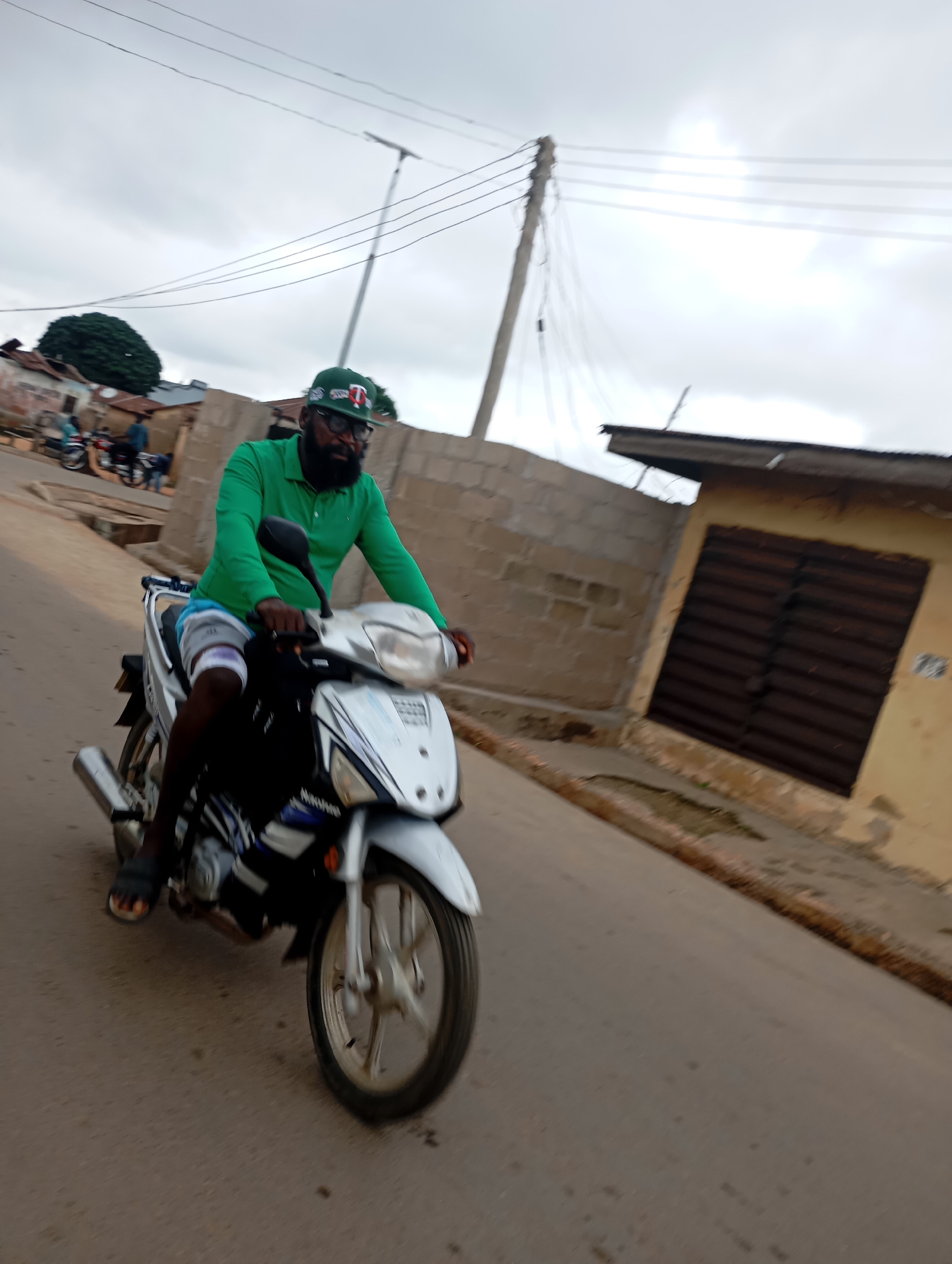 A man wearing a green polo shirt, blue and white patterned shorts, and a green baseball cap with a white logo is riding a silver and black motorcycle down a paved street in Shendam, Nigeria. He has a beard and is wearing glasses. The motorcycle is in motion, as indicated by the blur. The background shows buildings made of concrete blocks and corrugated metal roofs, with some trees and utility poles visible. The sky is overcast.