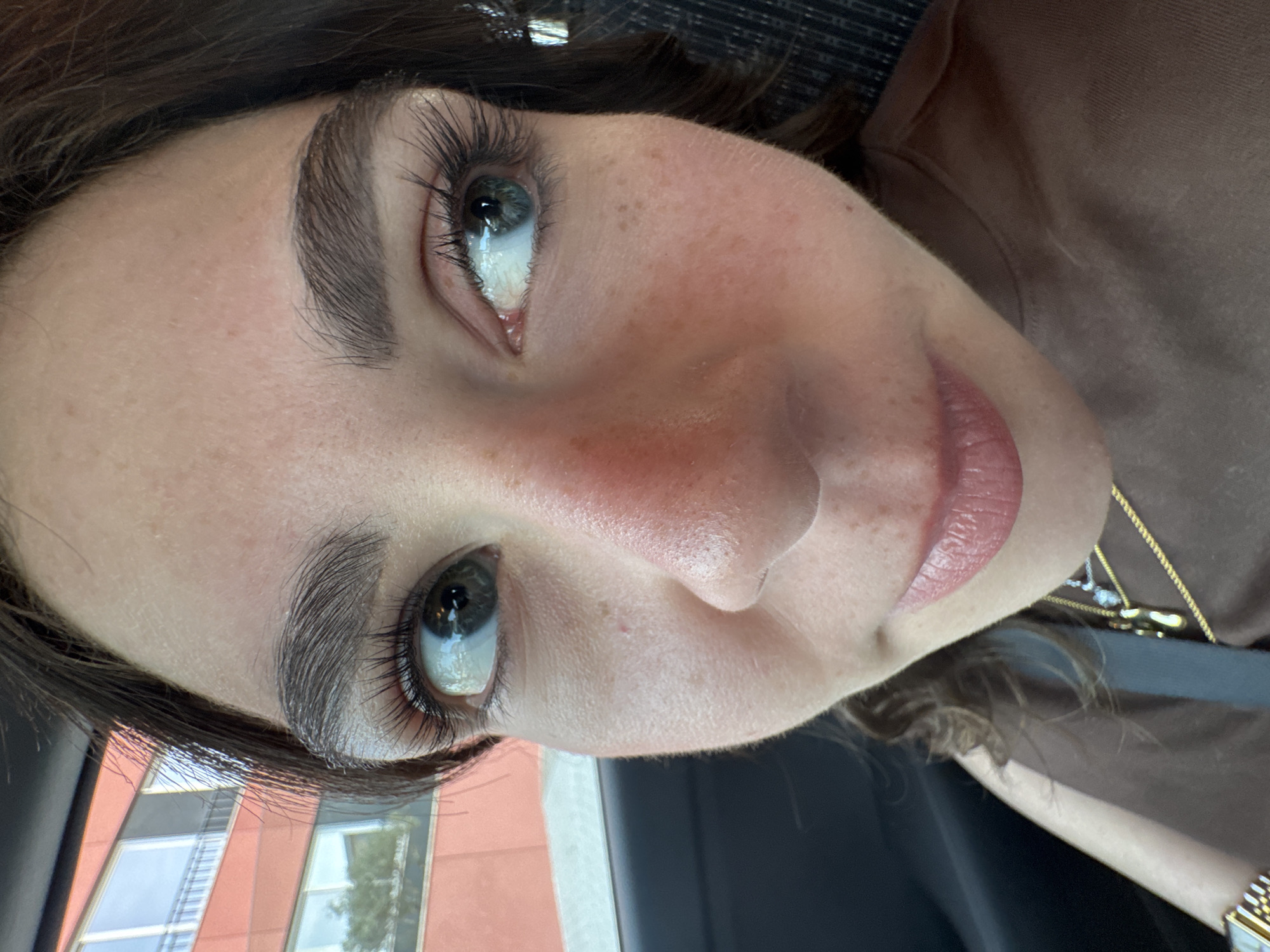A close-up, high-angle selfie of a woman's face. She has light skin with freckles, dark eyebrows, and long eyelashes. Her eyes are looking upwards and to the right. Her lips are slightly parted, revealing a light pink color. She is wearing a brown shirt, and a gold necklace is visible. Outside the car window, a building with a red and beige facade can be seen. The location cannot be verified in Mirandela, Portugal based on the provided information.