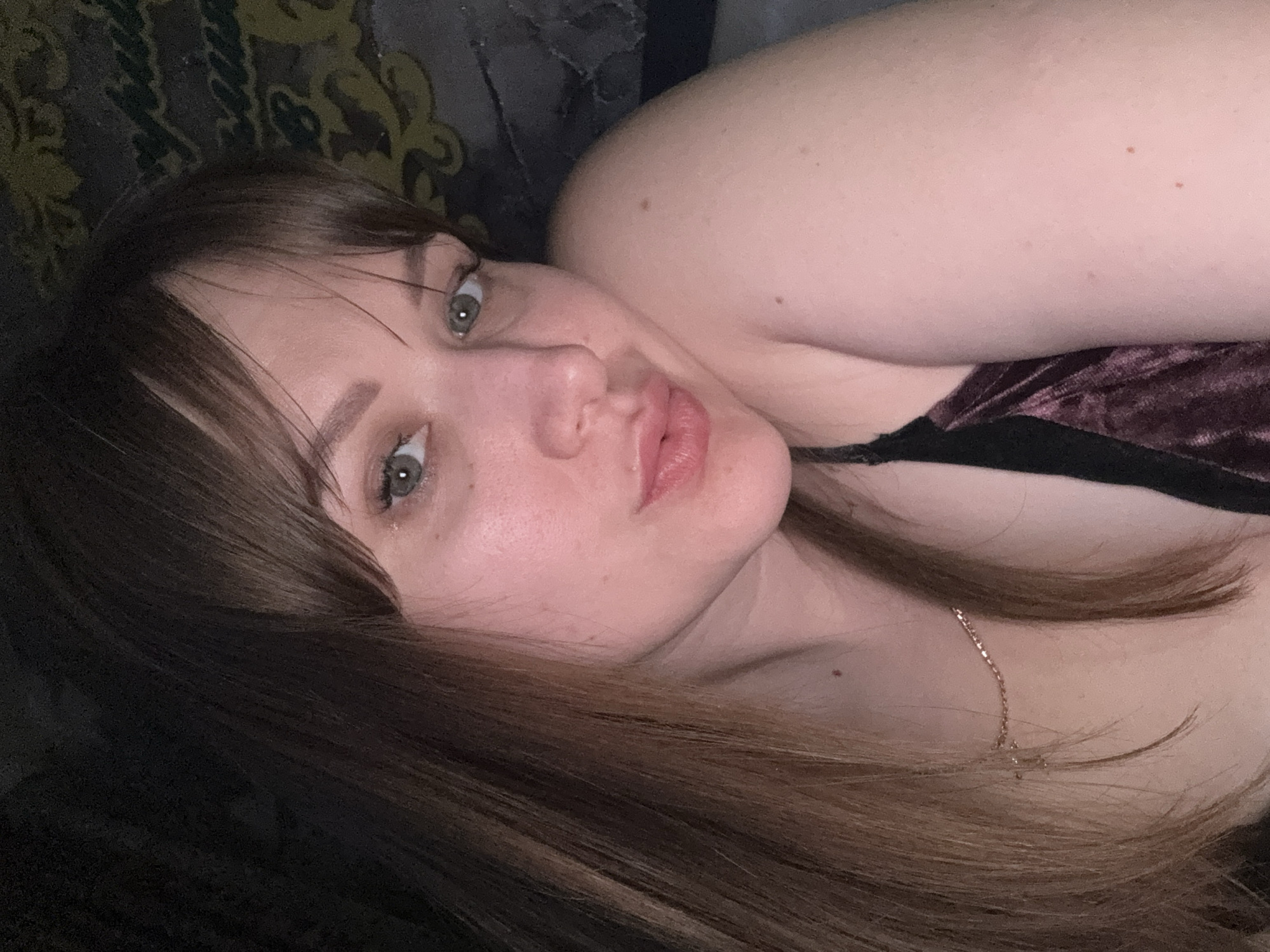 A woman with brown hair and green eyes is lying down. She is wearing a velvet top with black straps and a gold necklace. The background is dark and indistinct, possibly a patterned blanket or couch. The location cannot be determined from the image.
