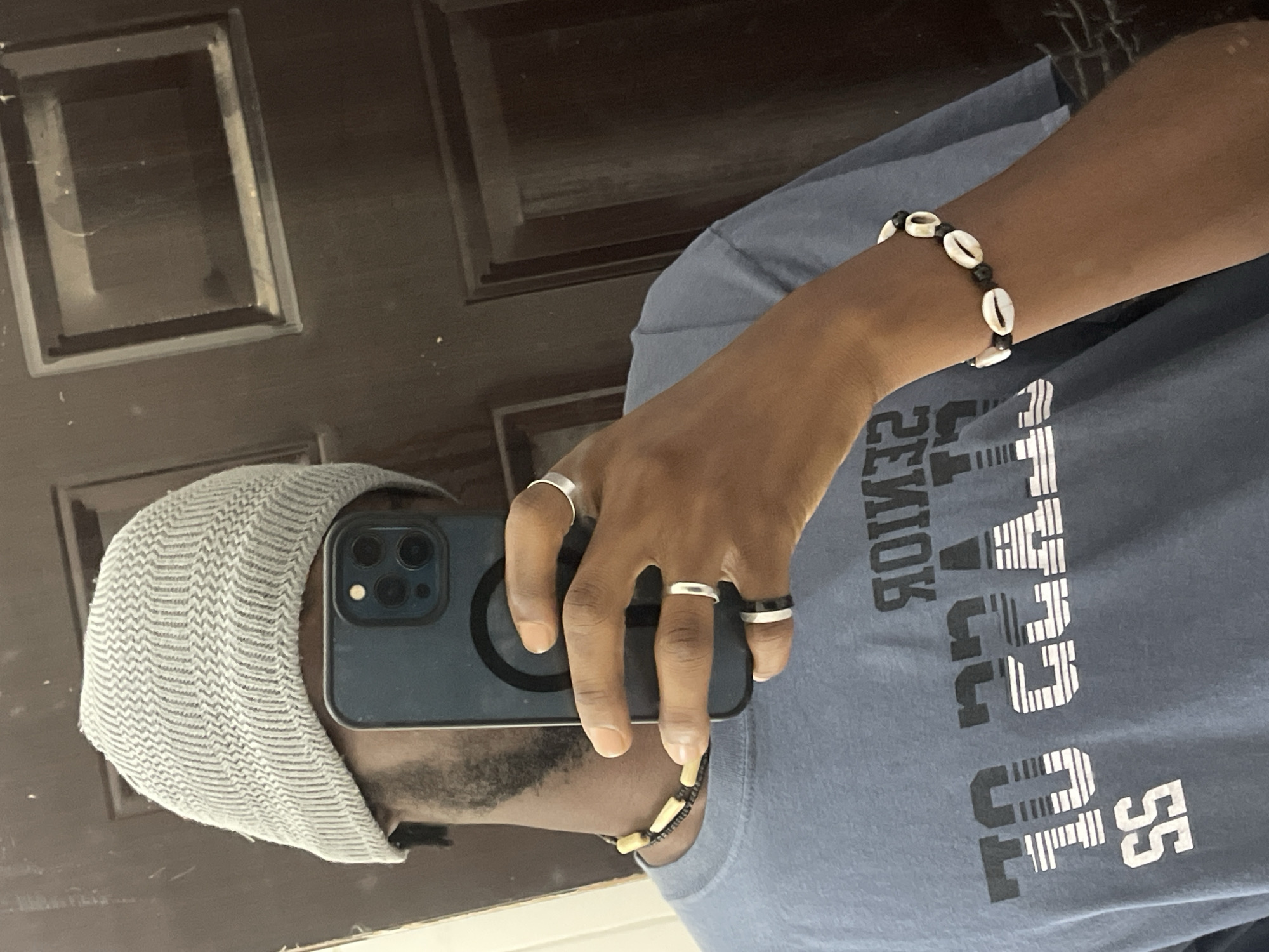 A man wearing a gray cap, a blue t-shirt with white and black text, and several rings is holding a smartphone and taking a selfie in front of a dark brown door. He is wearing a bracelet with white shells and dark beads on his right wrist and a necklace with brown and black beads. The location is not discernible from the image.