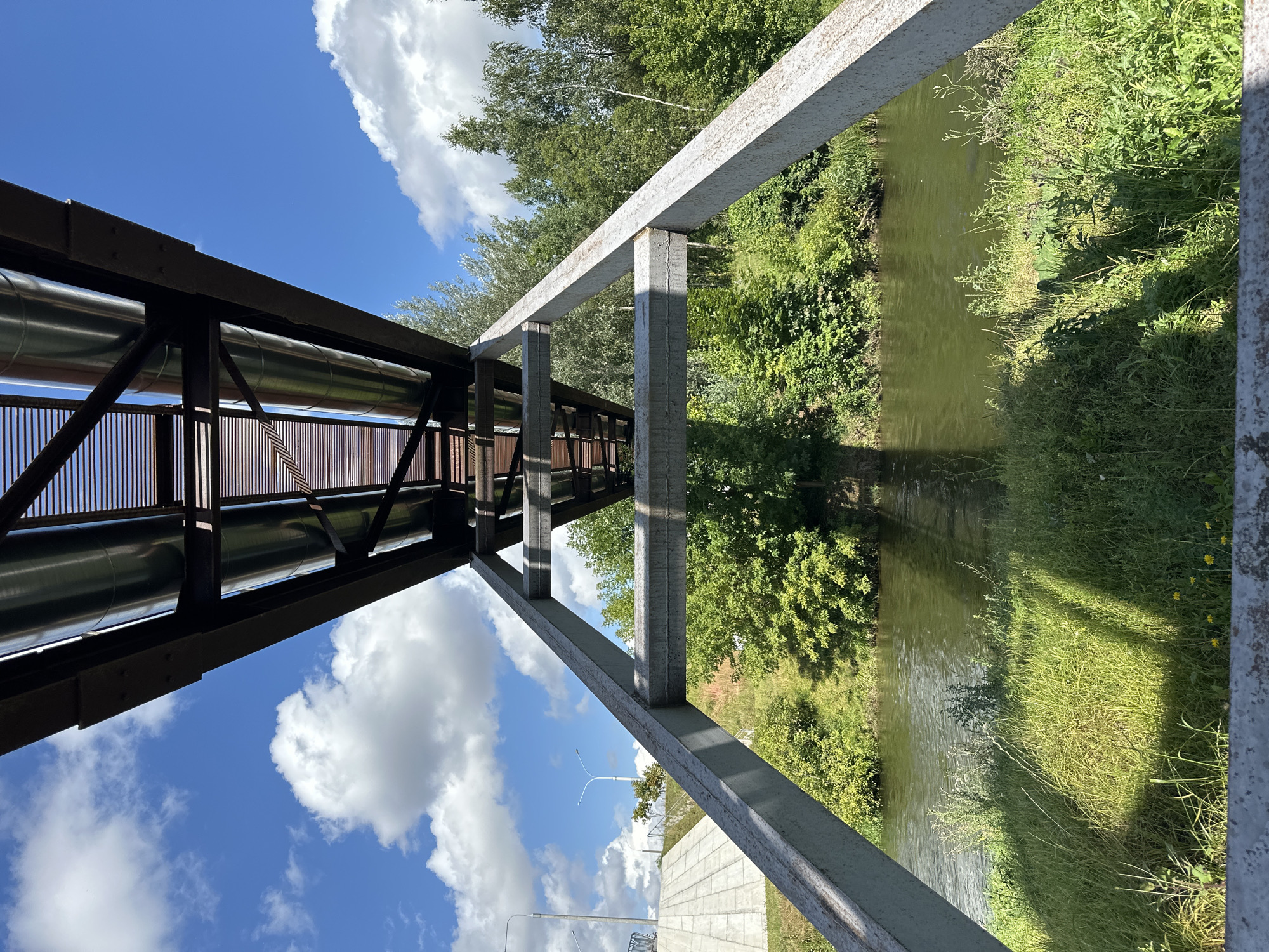This image, taken in the city of Minsk, Belarus, depicts an overhead view of large pipes supported by a metal structure, spanning across a calm body of water. The pipes are silver-colored and run parallel to each other, supported by a dark metal framework. The structure is propped up by two large, angled gray metal beams that frame the scene from the bottom.
The water below is a murky green, with gentle ripples indicating a slight current. The banks of the water are lined with lush green grass and dense vegetation. Sunlight casts shadows from the overhead pipes and beams onto the grassy areas, creating distinct patterns. In the background, a line of trees with dense green foliage stretches across the landscape, suggesting a park or natural area.
The sky above is a vibrant blue, dotted with fluffy white clouds. On the left side of the image, a concrete structure, possibly a ramp or embankment, is visible with streetlights in the distance. The overall impression is a utilitarian structure integrated into a natural setting. There are no people visible in the image.