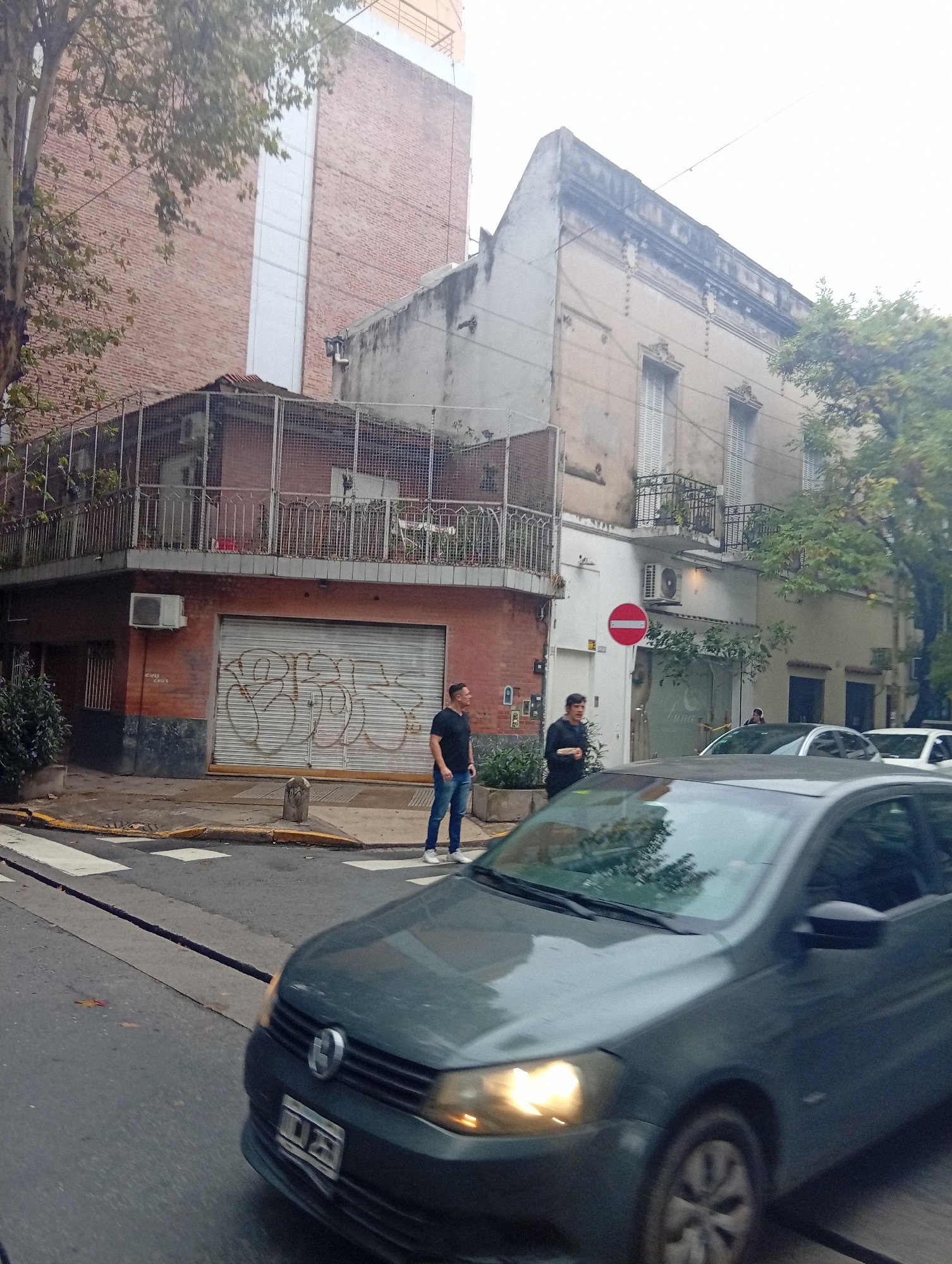 This image captures a bustling street corner in Buenos Aires, Argentina, under an overcast sky, likely during late afternoon or early morning, given the car's headlights.
In the immediate foreground, a dark grey Volkswagen Gol car is seen in motion, with its headlights on and its Argentine license plate partially visible on the front bumper. The street features a pedestrian crosswalk and what appears to be a linear track or groove running along the asphalt.
On the sidewalk, two men are visible. One man, wearing a black t-shirt and jeans, stands looking left towards the street. The other, wearing a dark jacket, is also observing the street scene.
The architecture in the background is a mix of styles. To the left, a multi-story building with red brick walls and a distinctive vertical white panel rises. Below it, at street level, is a commercial space with a grey corrugated metal security door covered in extensive white graffiti. Above this, a balcony enclosed by a metal cage-like railing wraps around the corner, holding plants and an air conditioning unit.
Adjacent to this, an older, cream-colored two-story building stands, featuring ornate decorative elements above its windows and balconies with dark metal railings. At its street level, another commercial establishment has a glass door with the word "Luna" visible in white text, and a red circular "No Entry" sign prominently displayed next to it. Another air conditioning unit is mounted on this building. Various trees with green foliage are scattered throughout the scene. Further down the street, other vehicles are partially visible.