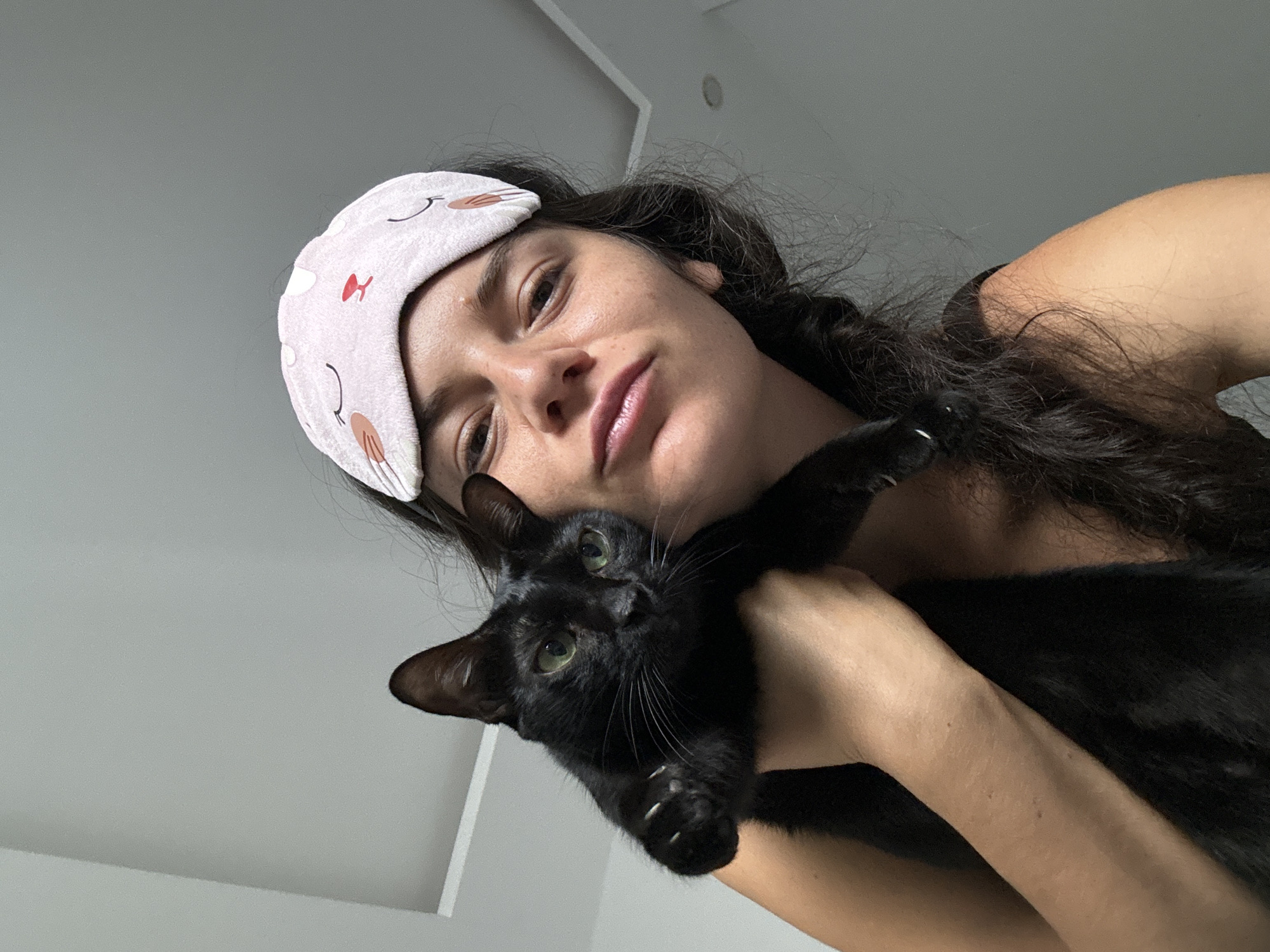 A close-up, low-angle shot features a woman with dark hair, wearing a light pink sleep mask with a cartoon cat face design, holding a black cat against her chest. The woman's eyes are open and looking towards the camera, and her lips are slightly parted. The cat has green eyes and its claws are visible on its outstretched paw. The background consists of a plain, light-colored ceiling with subtle linear detailing and a plain gray wall. The scene appears to be indoors. There is no indication of the city of Mũi Né, Vietnam.