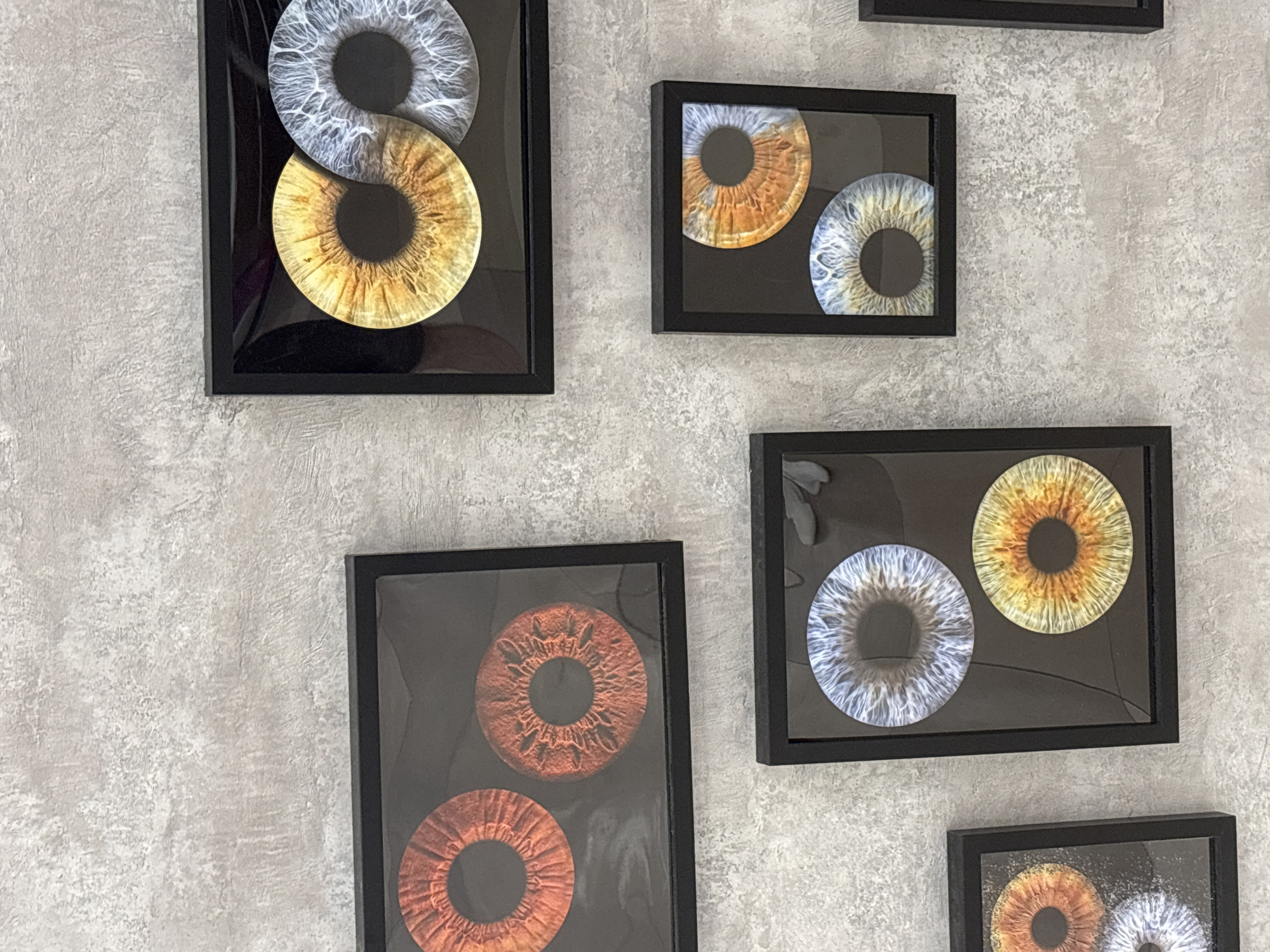The image displays a textured, light grey wall adorned with several black-framed artworks. Each frame showcases highly detailed, macro photographs of human irises set against a dark background.
In the top left, a large vertical frame features two overlapping irises: one is a light blue-grey with intricate white patterns, and the other is a warm golden-brown with radiating lines. To its right, a smaller horizontal frame contains a partial orange-brown iris and a complete light blue-grey iris.
Further down and to the left, a tall vertical frame, partially visible, holds two reddish-brown or copper-colored irises, one positioned above the other. Centrally located on the right side of the image, a medium-sized horizontal frame prominently displays a light blue-grey iris next to a bright golden-yellow one. A small, partially visible frame at the bottom right shows an orange-brown iris alongside a light blue-grey iris.
The wall itself has a rough, stucco-like or concrete texture. No people or activities are visible in the scene, which appears to be a static interior art display. There is no visible text or specific indication of the time of day or external location.