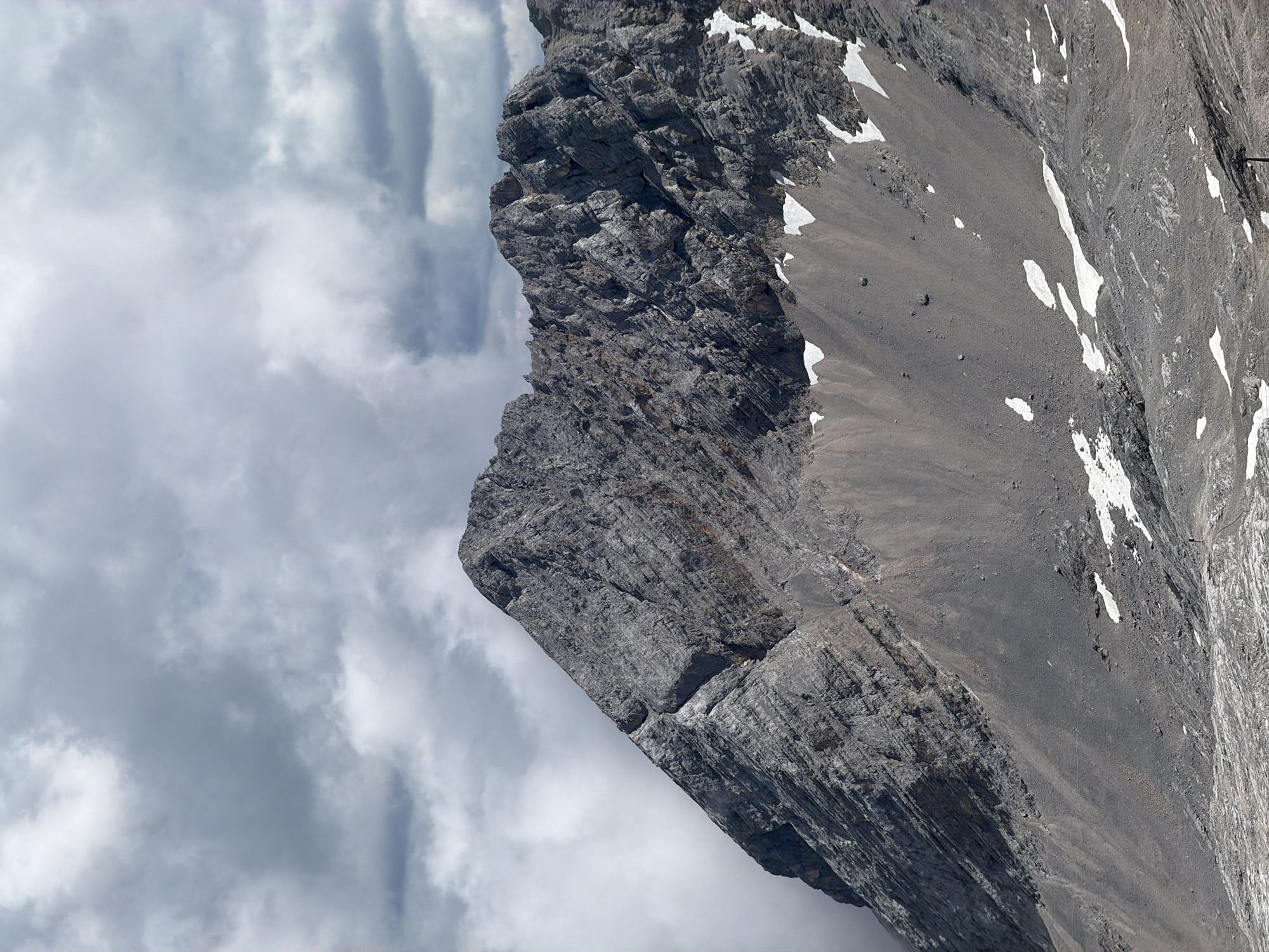 This image captures a close-up view of the Zugspitze, Germany's highest peak, located near Garmisch-Partenkirchen. The mountain is characterized by its rugged, rocky slopes, with visible layers and fissures in the stone. Patches of snow dot the landscape, contrasting with the grey and brown hues of the terrain. The sky above is filled with dramatic, dark clouds, suggesting a moody or overcast day. The foreground shows a rocky, uneven surface, potentially a glacier or scree slope, with a single ski lift pole visible in the lower right corner. The overall impression is one of a majestic and imposing natural landscape.