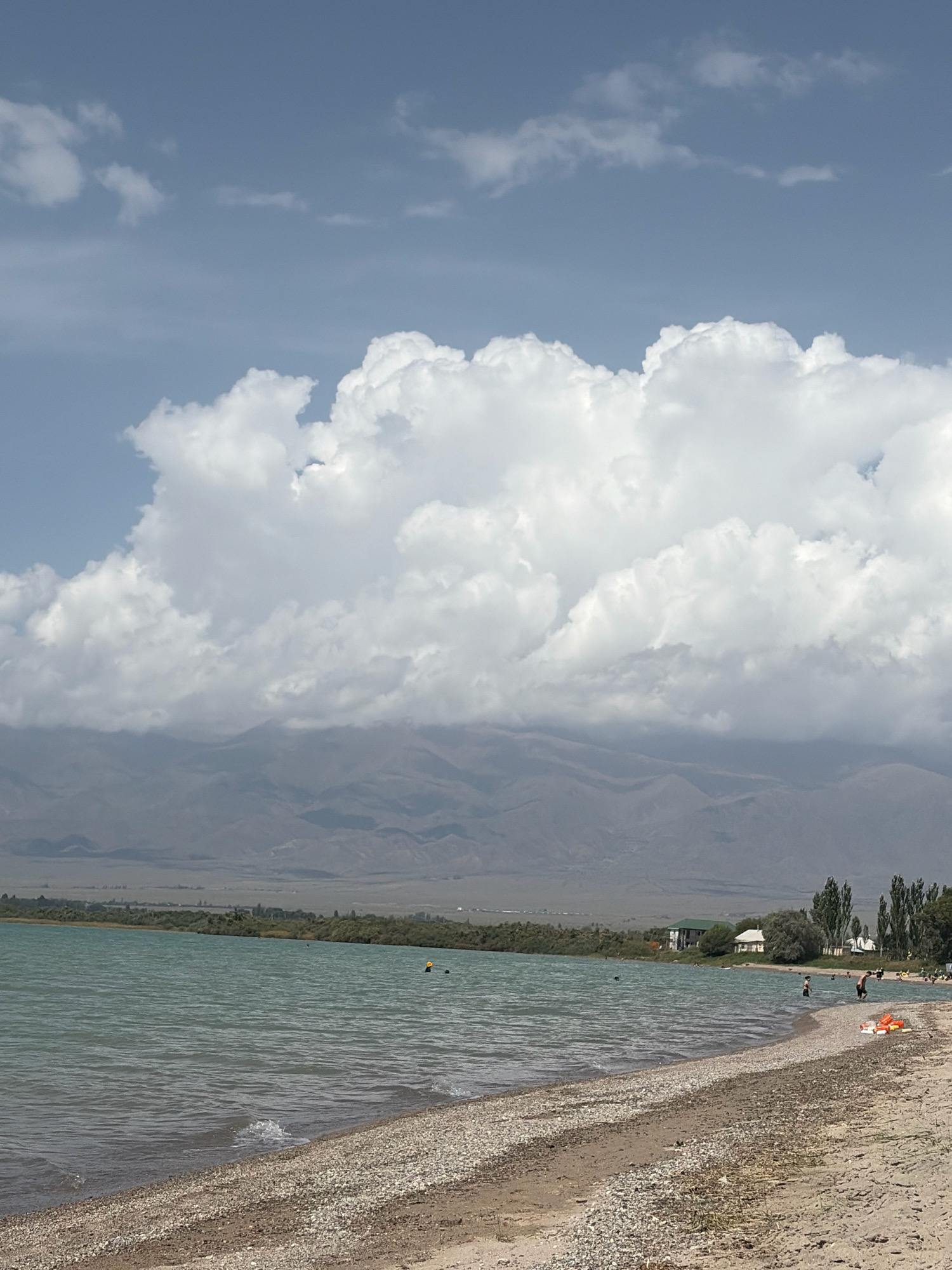 The image depicts a serene lakeside scene with a pebble beach in the foreground, leading to calm, turquoise water. In the distance, a line of trees and some buildings form the shoreline, with hazy mountains serving as a backdrop. The sky is a mix of blue with large, fluffy white clouds. Several individuals are visible near the water's edge, some swimming, others standing on the beach, and a few children are playing. There are also inflatable toys on the beach, suggesting a recreational area. The overall impression is one of a peaceful summer day at a lake.