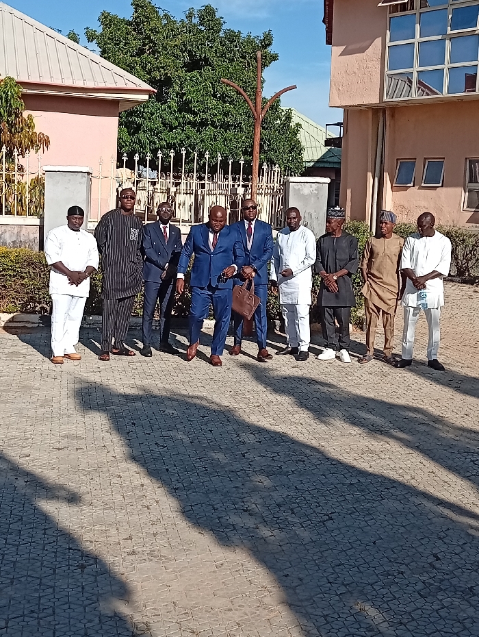 A group of nine men stands on a paved area. Some are in traditional attire, including white robes and hats. Others wear suits. A man in a blue suit is mid-stride. A few hold bags. Buildings are visible in the background, along with trees and a metal fence. Strong shadows are cast across the paved ground.