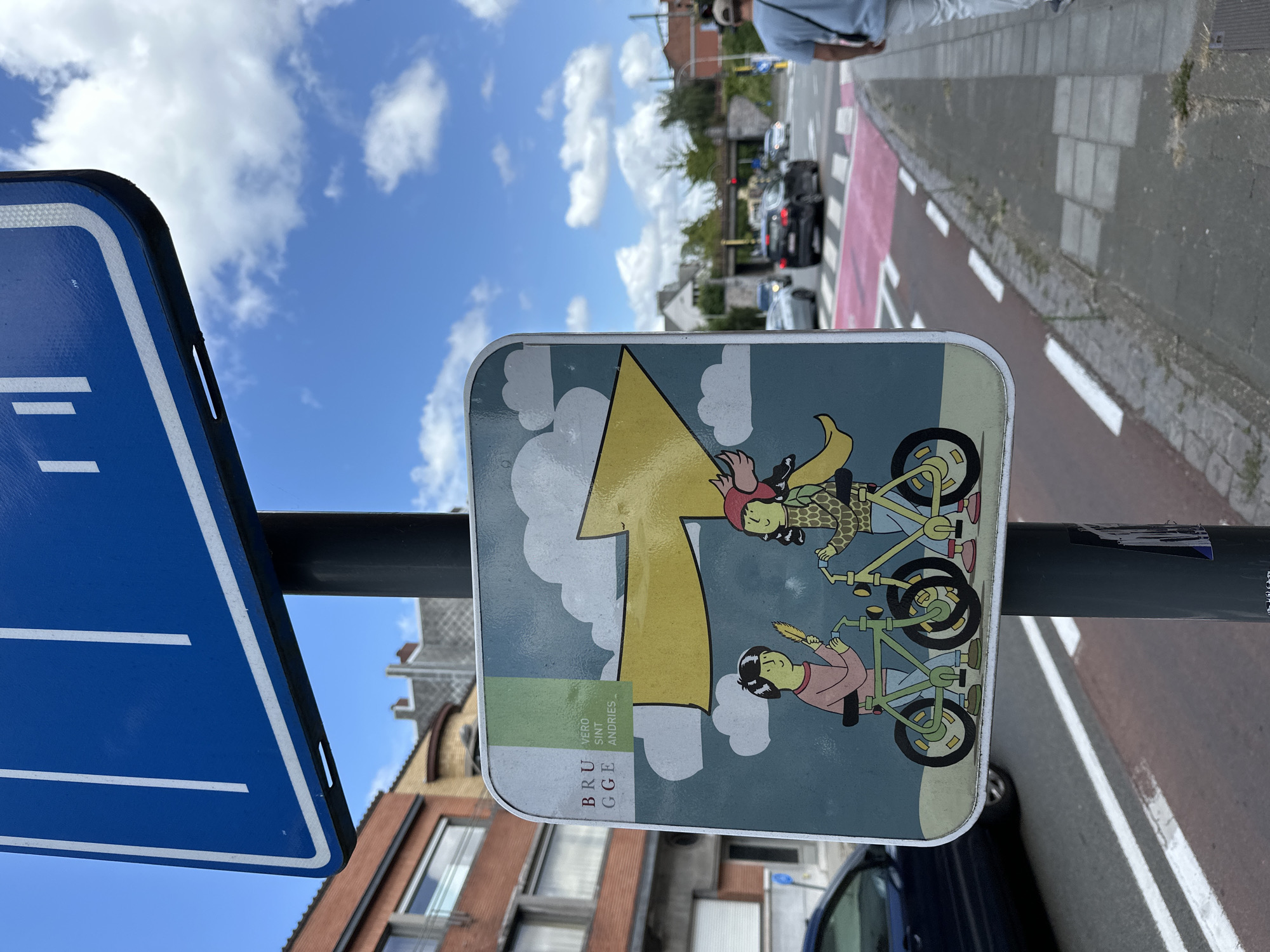A street sign with a cartoon illustration of two people on bicycles is mounted on a pole. The illustration shows two figures, one on the left with black hair wearing a pink shirt and holding a yellow feather, and the figure on the right with dark hair and a red hat wearing a patterned sweater and a yellow cape. They are both riding green bicycles. A large yellow arrow points to the right, above the cyclists, against a sky blue background with white clouds. To the left of the illustration, text reads "BRUGGE" and "VERO SINT ANDRIES." The sign is situated on a street with a red and white striped pedestrian crossing and a pink-colored bike lane. Several cars are visible in the background, driving on the road. The scene appears to be in Bruges, Belgium, given the text on the sign. The sky is blue with scattered white clouds, and buildings are visible in the background. A person is walking away from the camera on the right side of the image.