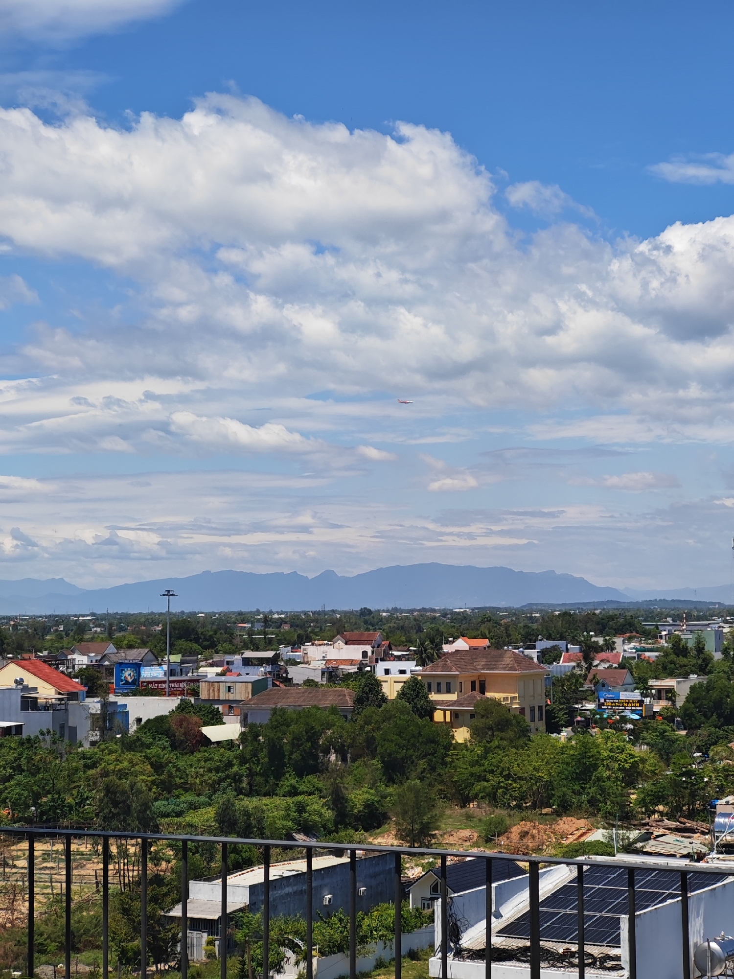 The image captures an aerial view of a city in Da Nang, Vietnam, under a partly cloudy blue sky. The scene showcases a dense residential area interspersed with lush green trees. Various houses, some with red tile roofs and others with modern designs, are visible. In the background, a range of mountains forms a distant silhouette against the horizon. A notable feature in the sky is a small red airplane, possibly a toy kite or a distant aircraft, adding a dynamic element to the otherwise static landscape. The foreground is partially occupied by a dark metal railing, suggesting the vantage point is from a balcony or elevated structure. Some buildings in the foreground appear to have solar panels installed on their roofs. The overall impression is that of a vibrant, developing urban environment with a strong presence of nature.