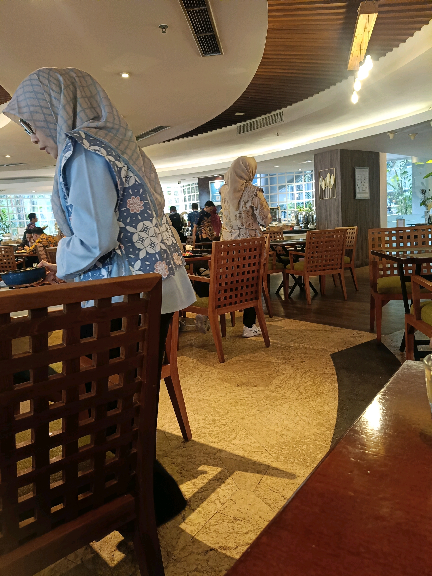 The image depicts the interior of a restaurant in North Jakarta, Indonesia. In the foreground, a woman wearing a blue hijab and a light blue shirt with a patterned vest is visible from the side, with her head tilted downwards as if looking at something on a table. She is partially obscured by a wooden chair with a lattice back. Further in the background, another woman in a light-colored hijab and a patterned dress is standing. Several other individuals, likely patrons and staff, are scattered throughout the restaurant, with some seated at tables and others standing. The environment appears to be a dining area with wooden tables and chairs, ambient lighting from ceiling fixtures and natural light coming from large windows in the background. The flooring is a textured pattern in shades of beige and brown.