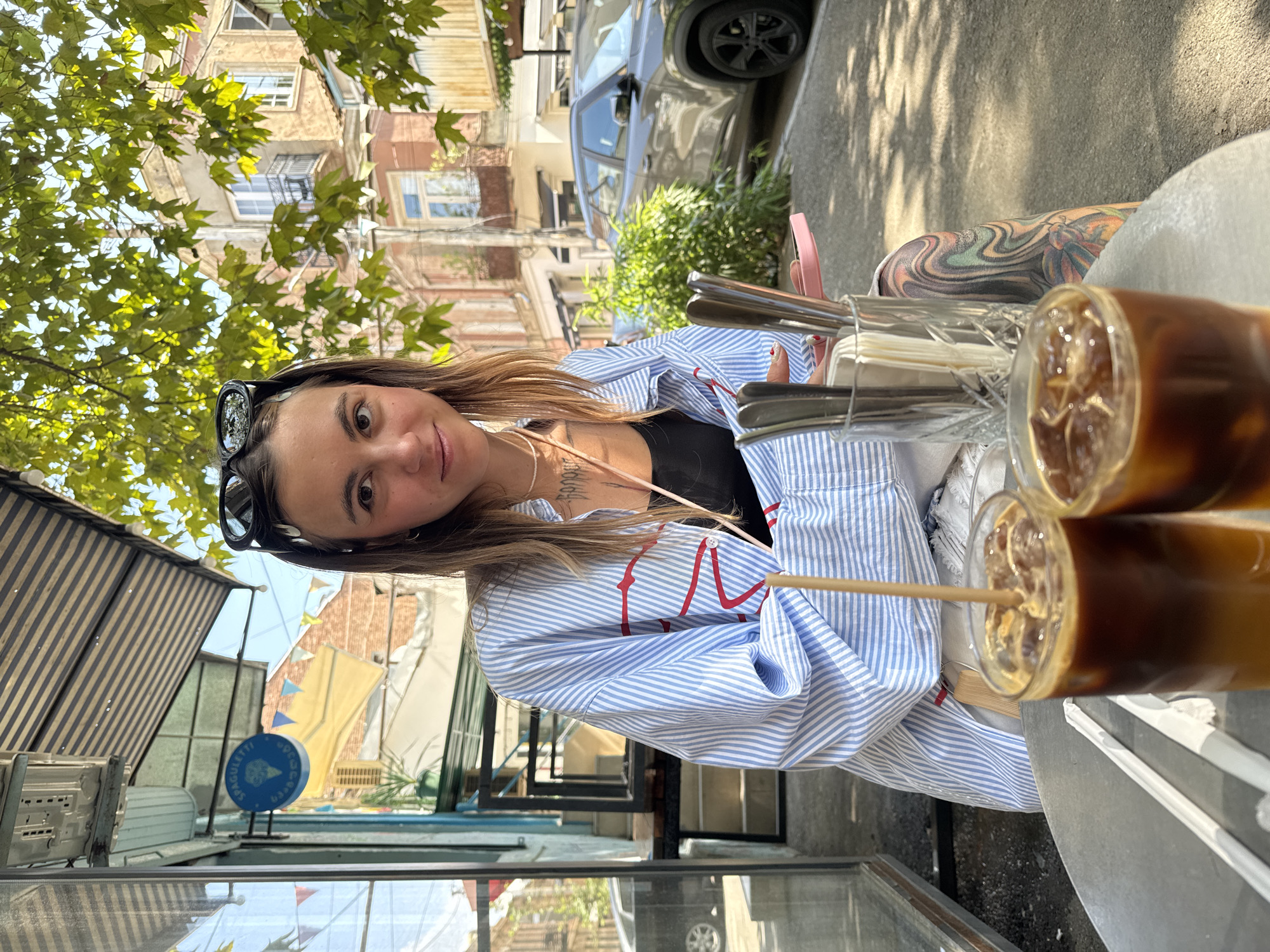 A young Caucasian woman with brown hair, wearing sunglasses perched on her head and a blue and white striped open shirt over a black tank top, sits at a metal table outside a cafe. She is looking towards the camera with a slight smile. In front of her are two glasses of iced coffee. A vase filled with cutlery sits on the table. The cafe appears to be located on a street in Tbilisi, Georgia, with buildings and trees visible in the background. A dark SUV is parked on the street behind the woman. The woman has tattoos on her arms and legs.