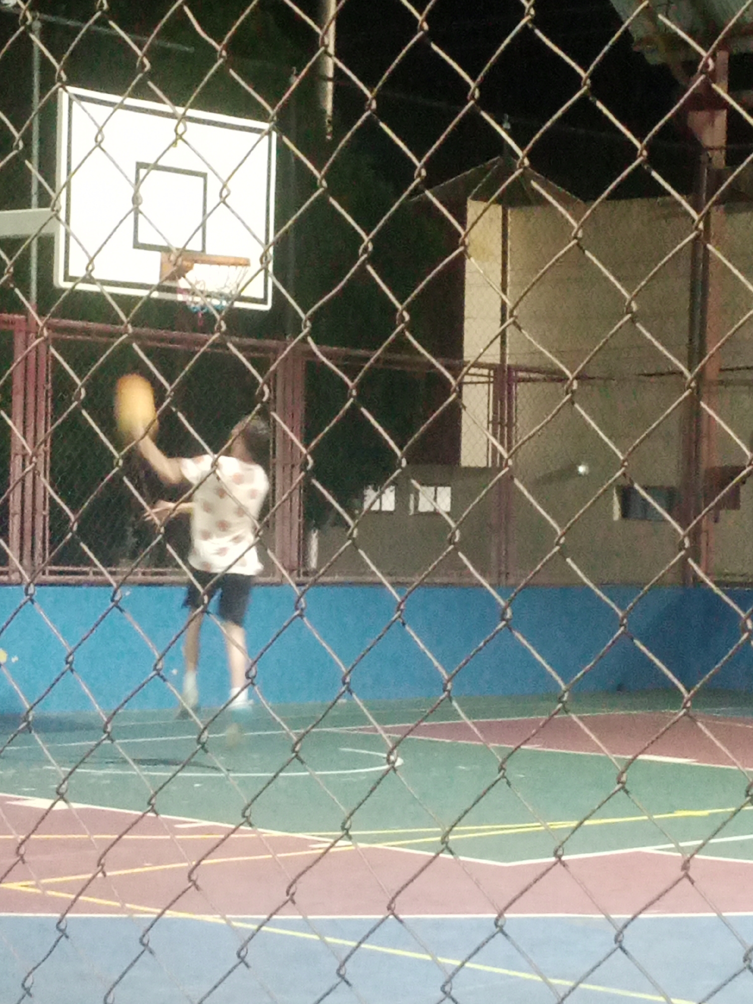 A person is playing basketball on an outdoor court in Santa Ana, Venezuela. The person, wearing a white shirt with a floral pattern and dark shorts, is mid-action with a yellow basketball in their left hand, raised as if preparing to shoot. The basketball court has a blue wall in the background and is marked with red, green, and yellow lines. A chain-link fence runs across the foreground, obscuring parts of the scene. The lighting suggests it is dusk or nighttime, with artificial lights visible in the background.