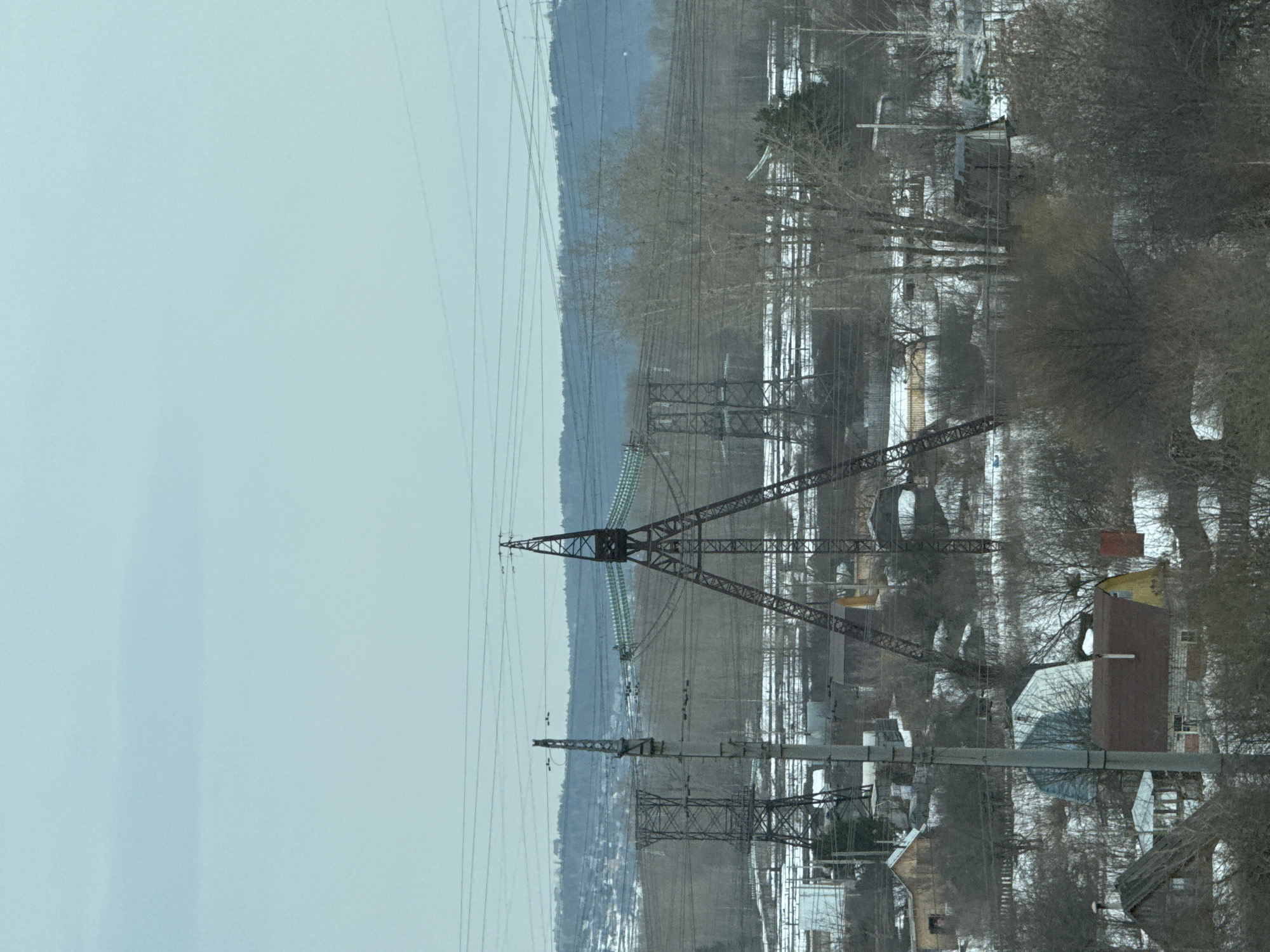 The image depicts a winter scene with a focus on power lines and infrastructure in a settled area. The location is Chaikovsky, Russia.
The sky is overcast. A series of large metal power line towers traverse the landscape, running from the horizon towards the viewer. Electrical wires are strung between the towers.
In the midground, there's a mix of trees, some bare due to the winter season. Scattered among the trees are houses and buildings, covered with snow. Roofs are visible.
The environment suggests a residential area on the outskirts of the city. The presence of power lines indicates access to electricity and communication infrastructure. There are no people, animals, or vehicles clearly visible in the image. The scene conveys a cold and quiet winter day in a semi-urban or rural setting of Chaikovsky, Russia.
