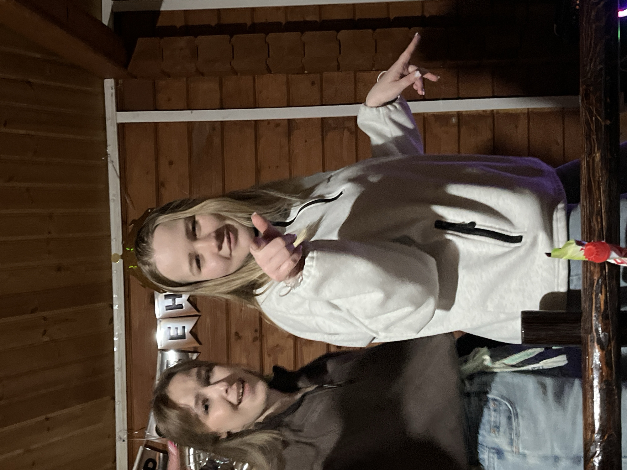 Two young women are posing for a photo. The woman on top is wearing a white hooded sweatshirt and has blonde hair. She is giving a thumbs-up with one hand and holding up two fingers with the other. The woman below is wearing a brown zip-up jacket. Behind them is a wooden wall, and part of a "HOME" balloon banner is visible. Based on the environment and previously mentioned location information, the photo was likely taken indoors, possibly at a private residence in Chaikovsky, Russia.