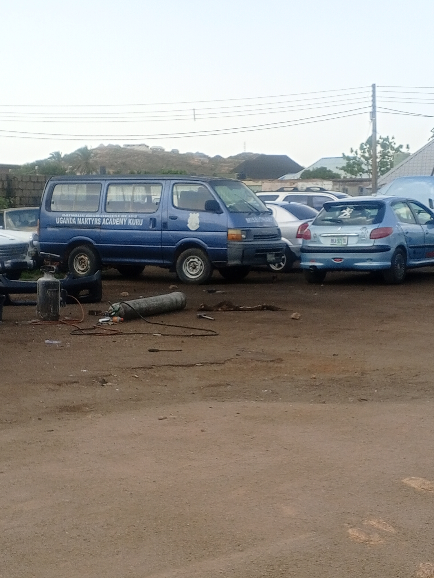 This media file captures an outdoor scene, likely a repair shop or service area, with several vehicles present. A prominent blue van, emblazoned with the text "CATHOLIC ARCHDIOCESE OF JOS UGANDA MARTYRS ACADEMY KURU," is parked in the foreground. Beside the van, two large gas cylinders are visible, one upright and another lying horizontally, suggesting repair or maintenance work is in progress. Several other cars, including a light blue Peugeot hatchback with a license plate that reads "MAK-92TX," are parked in the background, some partially obscured. The setting appears to be an unpaved area, with dirt and scattered debris on the ground. In the distance, a slightly elevated landscape with vegetation and structures is visible under a bright, overcast sky, indicating daytime. The overall mood is functional and utilitarian, with no explicit human presence or emotional cues. The presence of the gas cylinders and tools points towards an activity related to vehicle repair or welding.