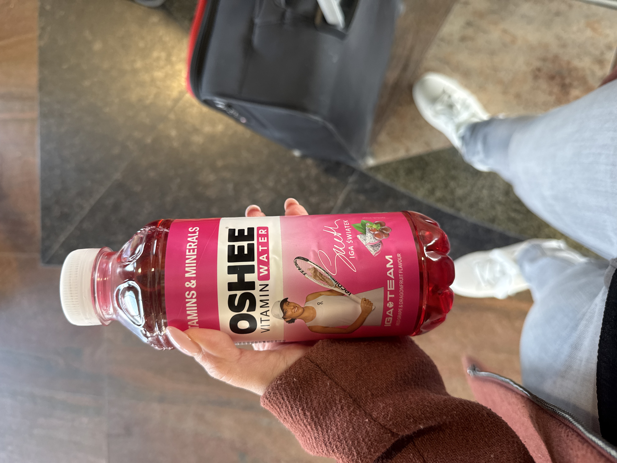 A person is holding a bottle of OSHEE vitamin water with Iga Świątek's image on it. The bottle is red grape and dragonfruit flavored. The person is wearing a brown sweater with a pink detail. In the background are light blue jeans, white shoes, and a red and grey suitcase. The floor appears to be tiled and patterned. There is no indication in the image that suggests this photo was taken in Sofia, Bulgaria.