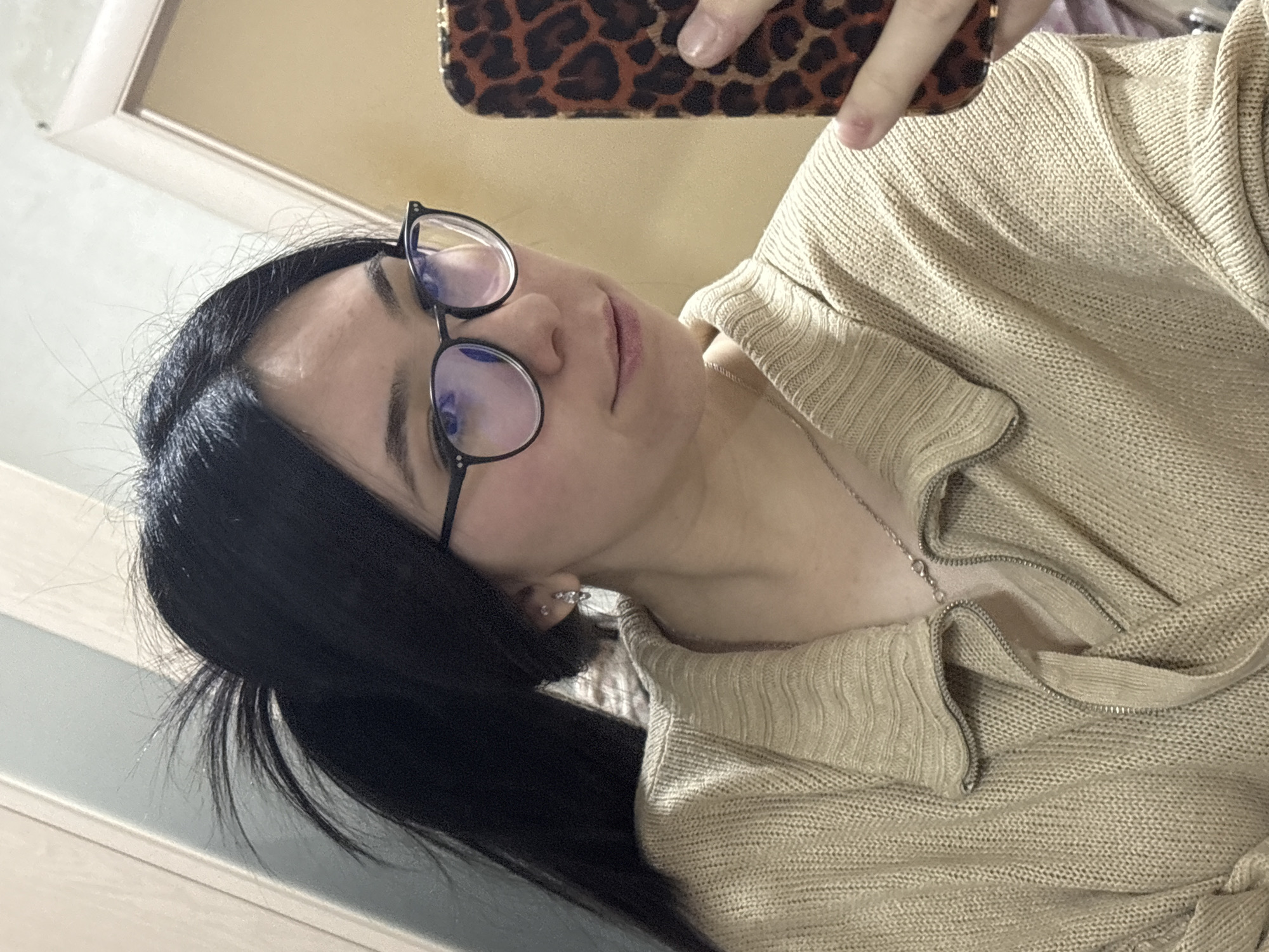 This close-up mirror selfie captures a young person, likely in Izhevsk, Russia. The subject, a female, has long, dark hair pulled back, and is wearing black-framed glasses with a slight bluish tint reflecting in the lenses. Her expression is neutral to slightly pensive, and her lips appear lightly colored.
She is dressed in a light-colored, possibly beige or pale yellow, knitted sweater or cardigan featuring a collared neckline and a silver-colored zipper. A delicate silver chain necklace is visible around her neck, and a small, reflective earring can be seen on her left earlobe. Her nails are painted a light, natural hue.
The selfie is taken with a smartphone, partially visible at the top, featuring a distinct leopard print case, and held by her hand. The image is oriented diagonally, suggesting the angle at which the photo was taken in the mirror. The background shows reflections of an indoor setting, including a light-colored ceiling or wall and what might be a door frame. No visible text is present in the image.